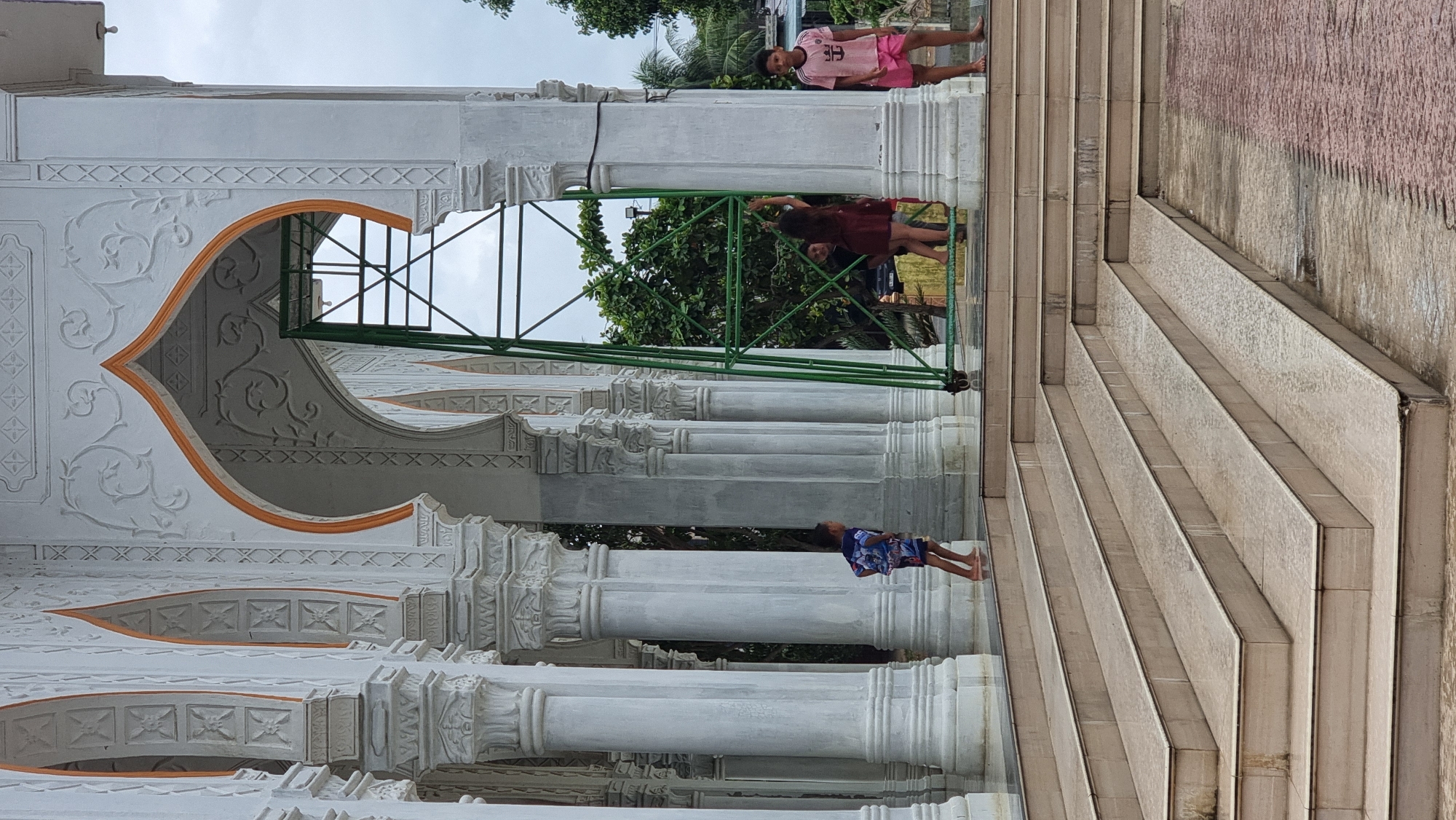 This image captures a scene at the Baiturrahman Mosque in Meunasah Lhok, Indonesia. In the foreground, a set of wet, tiled steps leads up to a covered walkway supported by ornate white columns. The walkway itself has a wet, reflective floor. A young boy, wearing blue and white shorts and a blue and white patterned shirt, stands on the walkway, facing left and looking away from the camera.
Behind the boy, the walkway continues, featuring a series of large white columns with intricate decorative carvings. An arched opening, highlighted with orange trim and also adorned with carvings, leads to an area where a green metal structure stands.
Further into the scene, several people are visible. A person in a maroon dress is interacting with the green structure. To their right, another person, possibly a child, is also near the structure. To the far right, a boy wearing pink shorts and a pink t-shirt stands on the walkway, facing towards the viewer. The overall environment appears to be outdoors, with trees and a cloudy sky visible in the background. The architecture suggests a mosque or similar religious building.