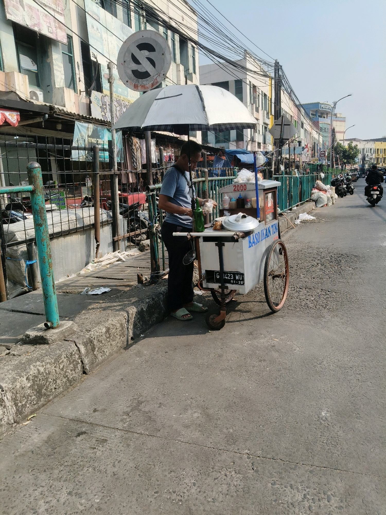 An outdoor, medium shot captures a street vendor's cart on a sunny day in South Tangerang, Indonesia. The vendor, a man wearing a blue t-shirt and dark pants, stands beside his cart, which is laden with food items and condiments. The cart is white with blue lettering that reads "BASO IKAN TUNA". A license plate is visible on the cart, reading "G 3423 WU 01-25". A white umbrella provides shade over the cart.
Behind the cart, a metal fence encloses what appears to be a walkway or a small channel. Various stalls and buildings line the street, many with signage and advertising. The street itself is paved and shows signs of wear. In the background, motorcycles are visible, some parked and others in motion, indicating a typical street scene in a bustling urban environment. The sky is clear and blue. The overall impression is of a vibrant, everyday scene on a city street.