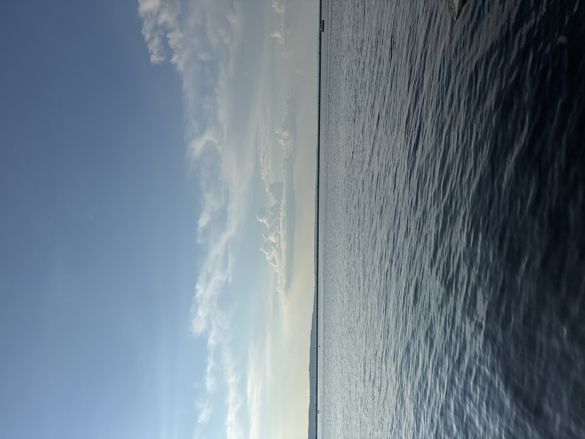 The image depicts a calm body of water with a long, low-lying shoreline in the distance. The sky is a clear blue with scattered white clouds, some of which are puffy and cumulus-like, while others are wispy cirrus clouds. There are several small boats visible on the water, and a line of structures, possibly poles or towers, stretches along the horizon. The water itself is a deep blue with gentle ripples, reflecting the sky. The scene appears to be a natural landscape, likely a reservoir or a large lake, given the expansive view of the water and the distant shoreline. The area is likely the Kama Reservoir, near Chaikovsky, Russia, a location known for its scenic water views and recreational activities. The image conveys a sense of tranquility and open space, typical of a summer day by the water.