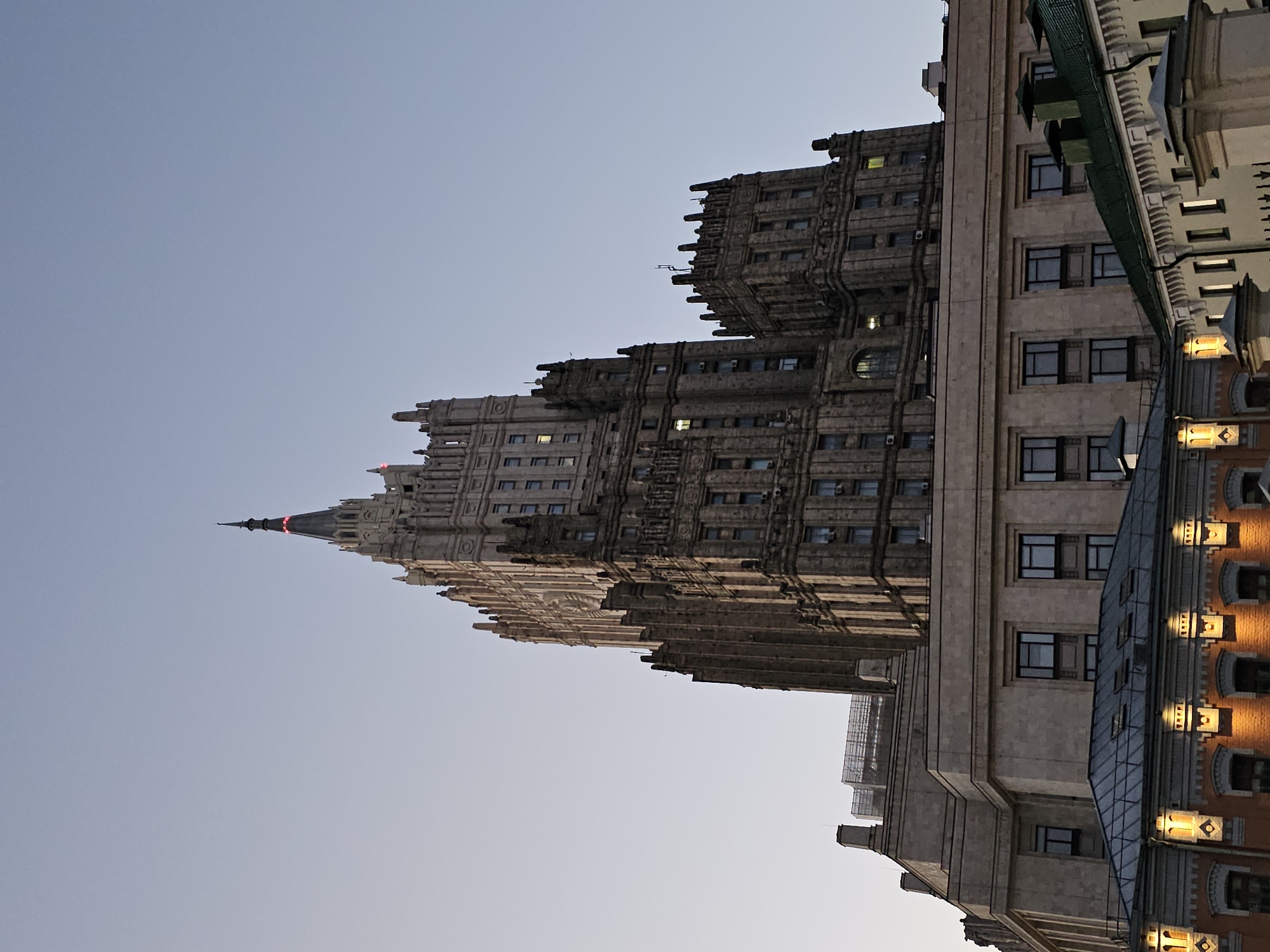 The image shows the Ministry of Foreign Affairs building in Moscow, Russia, at dusk. The iconic Stalinist skyscraper, with its distinctive spire topped with a red star, dominates the frame. The building is made of stone and has many windows. In the foreground, there are several other buildings with green roofs and illuminated windows. The sky is a pale blue, indicating the transition from day to night. There are no people visible in the image.