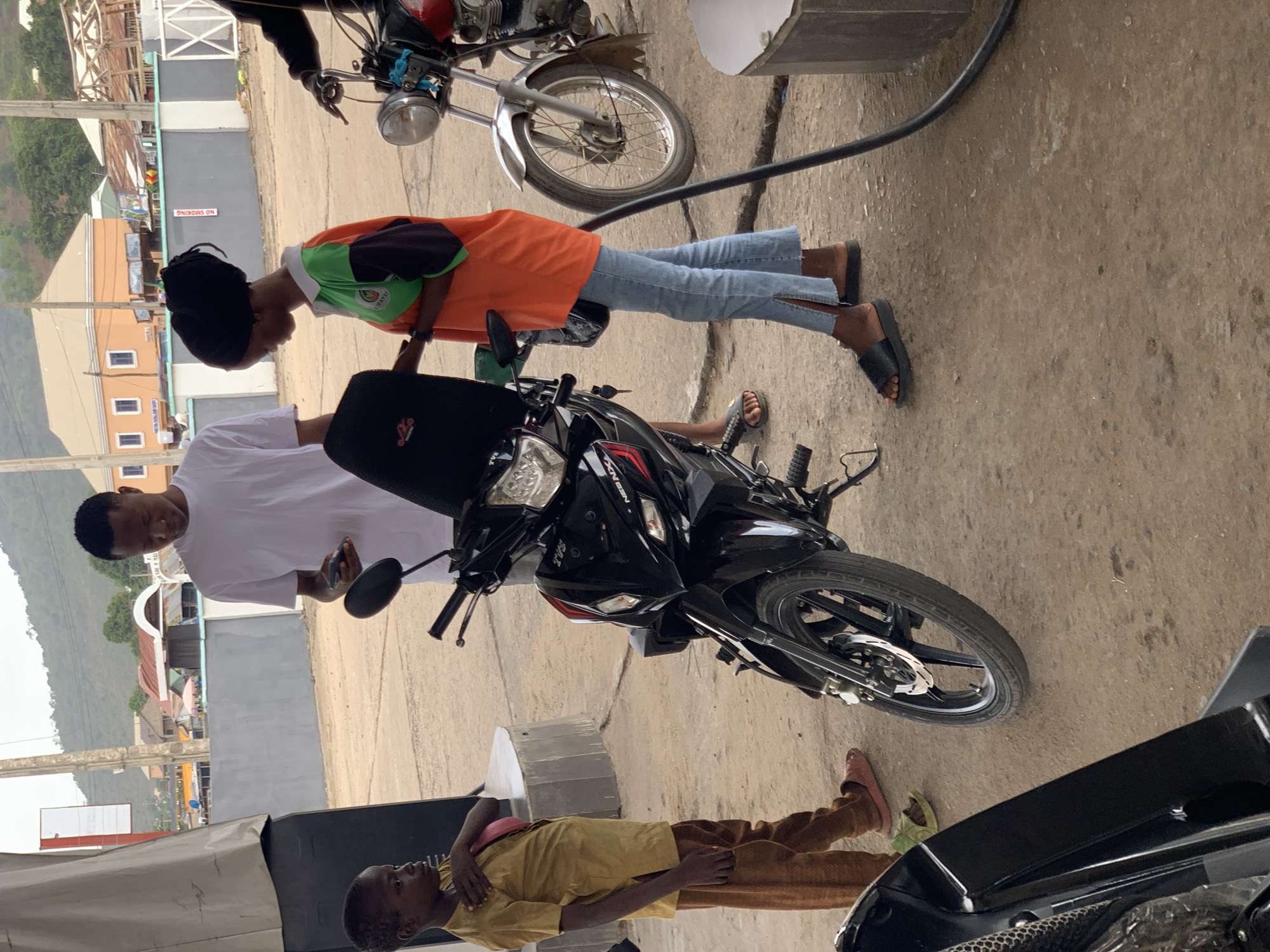 The image depicts a scene at what appears to be a petrol station in Lokoja, Nigeria. Two motorcycles are present. A person wearing an orange shirt and blue jeans is standing near one of the motorcycles, seemingly interacting with it. A man in a white shirt is standing to the left of the motorcycle. A younger person wearing a brown outfit is lying on the ground nearby. The background shows buildings, mountains and sky. A gas pump is also visible. The scene appears to be taking place outdoors.