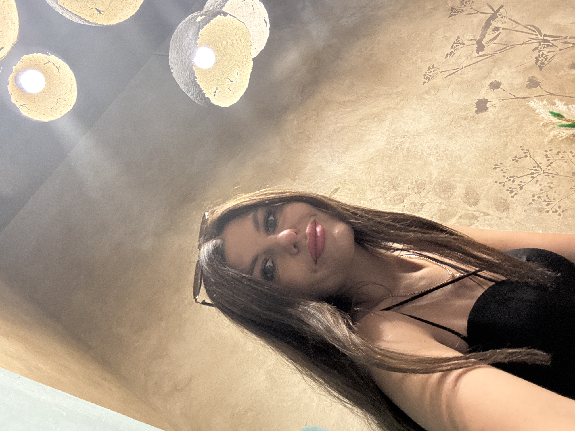 A woman with long, dark brown hair and light makeup is looking at the camera. She is wearing a black spaghetti strap top and a necklace. Sunglasses are resting on top of her head. She is positioned in the foreground, with a textured, light brown wall behind her. The wall has a subtle pattern of silhouetted plants. In the upper right corner, there are several spherical light fixtures with a rough texture, emitting a warm glow. The overall lighting suggests an indoor setting, possibly a restaurant or cafe. There are no other individuals visible in the frame. The image appears to be a selfie, taken from a low angle. The location is identified as Izhevsk, Russia.