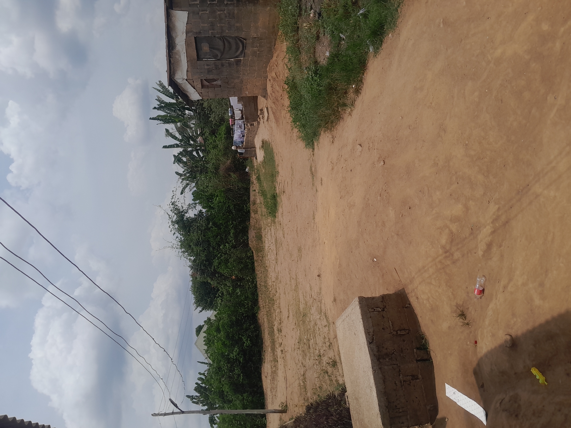 The scene, set in Ifo, Nigeria, depicts an unpaved reddish-brown dirt landscape under an overcast sky with scattered grey clouds. In the lower-left foreground, a low concrete block structure is visible. Adjacent to it on the dirt are scattered items: a red plastic bottle, a small red plastic cap, a piece of white paper, and a yellow plastic object. A slender utility pole stands on the mid-left, from which three dark power lines extend diagonally across the upper frame. To the right, a single-story building constructed of grey concrete blocks features a light-colored material along its roofline and a dark fabric covering a rectangular opening on its facade. A smaller, open window is present to the left of this covered opening. Behind this building, a lower concrete platform supports clothes drying on a line. Dense green vegetation, including bushes and banana trees, occupies the background and mid-ground left. No human figures are visible.