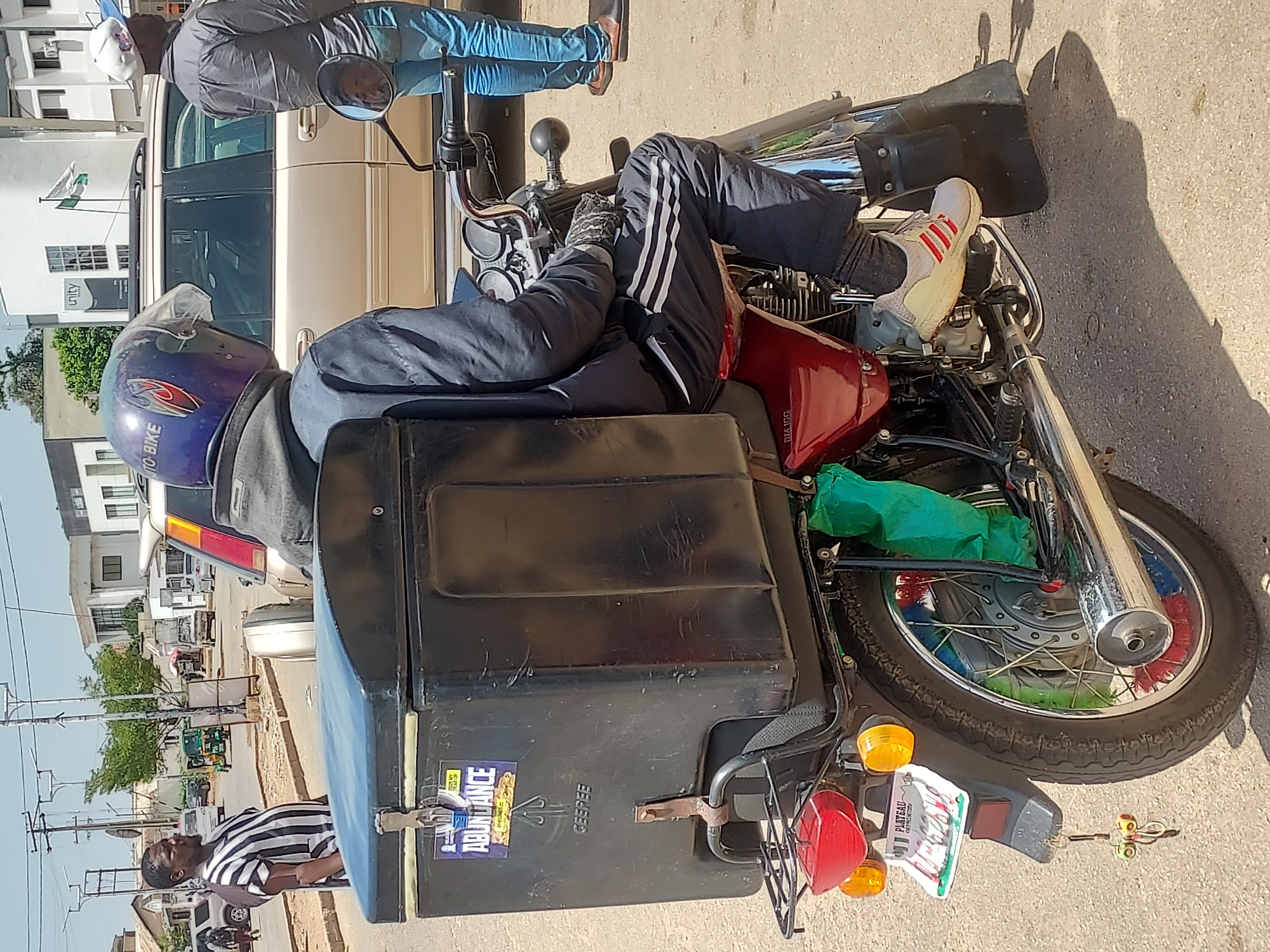 A man wearing a striped shirt stands next to a motorcycle. The motorcycle is equipped with a large black cargo box attached to the back. The rider is wearing a blue helmet, a grey jacket, black track pants with white stripes, and white Adidas sneakers. The motorcycle has a red fuel tank, and its front wheel has colorful decorations. The motorcycle has a "PL Plateau" license plate. A car is parked next to the motorcycle, and other buildings and vehicles are visible in the background. A sign reads "unity". The photograph was most likely taken in Jos, Nigeria.