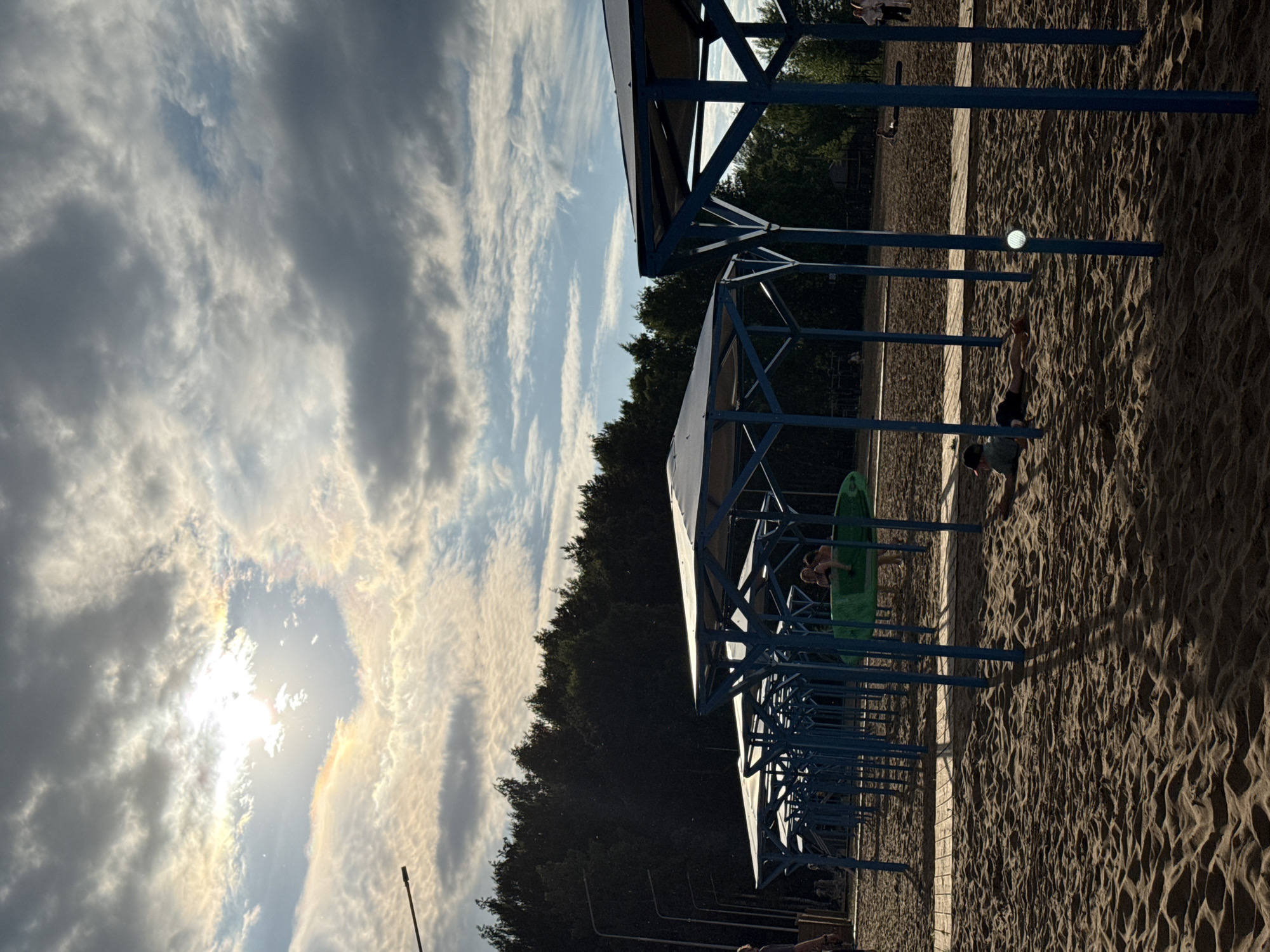 The image depicts an outdoor scene with a sandy ground, likely a beach or a recreational area. Multiple blue metal structures, possibly shelters or gazebos, are visible, casting long shadows on the sand. In the background, a dense line of dark green trees forms a backdrop against a sky filled with dramatic, wispy clouds. The sun is shining through the clouds, creating a bright, somewhat diffused light that suggests it is either early morning or late afternoon. The presence of people in the image indicates a recreational activity is taking place. One person is lying on the sand, and another is standing near a green object, possibly a mat or a surf-like board. The overall atmosphere appears relaxed and leisurely. The image is oriented vertically. There is no visible text in the image.