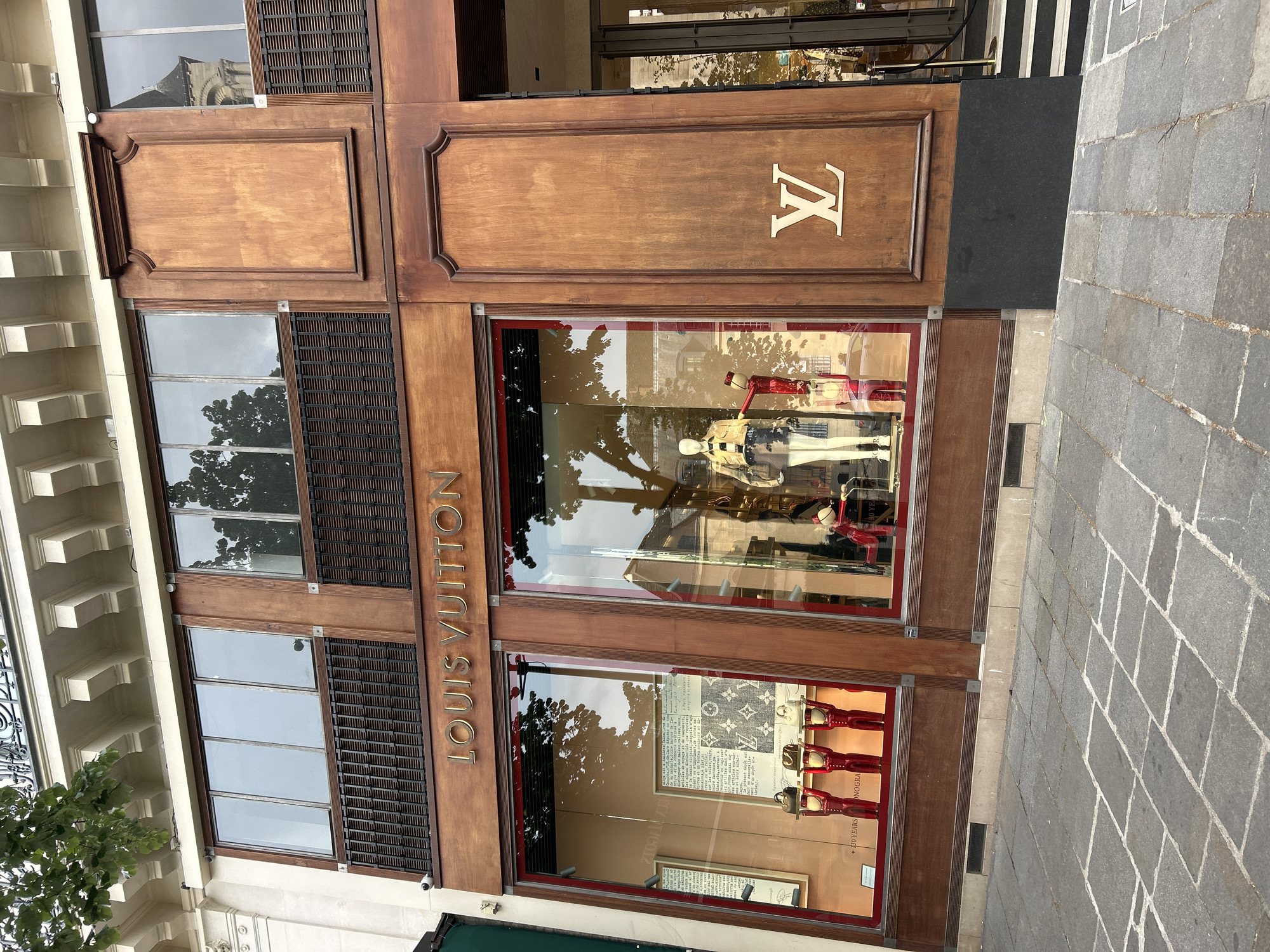 Here is a detailed description of the image:
**Scene:** The image shows the storefront of a Louis Vuitton store.
**Environment:** The store is located on a stone-paved street. The building exterior features wooden panels and window displays. Reflections of buildings and trees are visible in the windows.
**Objects:** Key objects include:
* The Louis Vuitton logo (both the monogram and the name signage)
* Window displays featuring mannequins wearing clothing and accessories.
* Displays of bags and promotional materials in the lower window.
**Details:** The wooden panels of the store are intricately designed with molding and a rich wood tone. The stone paving of the street appears well-worn.
**Inferences:** Based on the quality of the store and its brand, the location is likely an upscale shopping district in a major city. The reflection in the windows confirms an urban setting with trees.