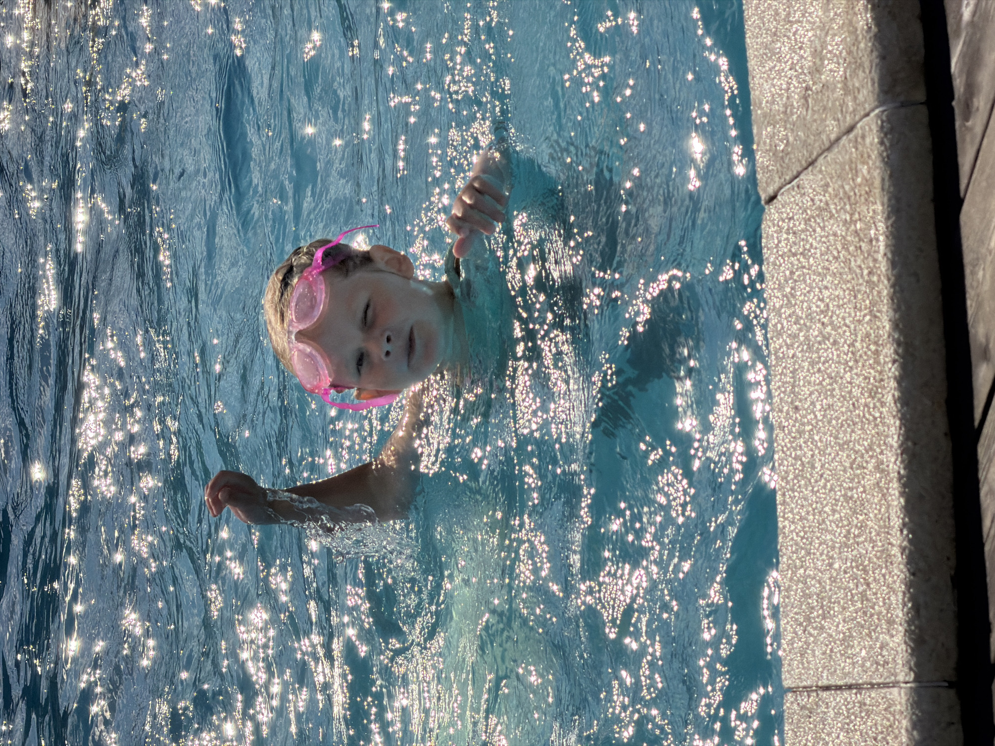 A young girl with pink goggles is swimming in a pool. She is looking at the camera and has her right arm raised out of the water. The water is blue and reflects the sunlight. The edge of the pool is visible in the foreground. The scene appears to be in a backyard pool, not in the city of Kamyshin, Russia.