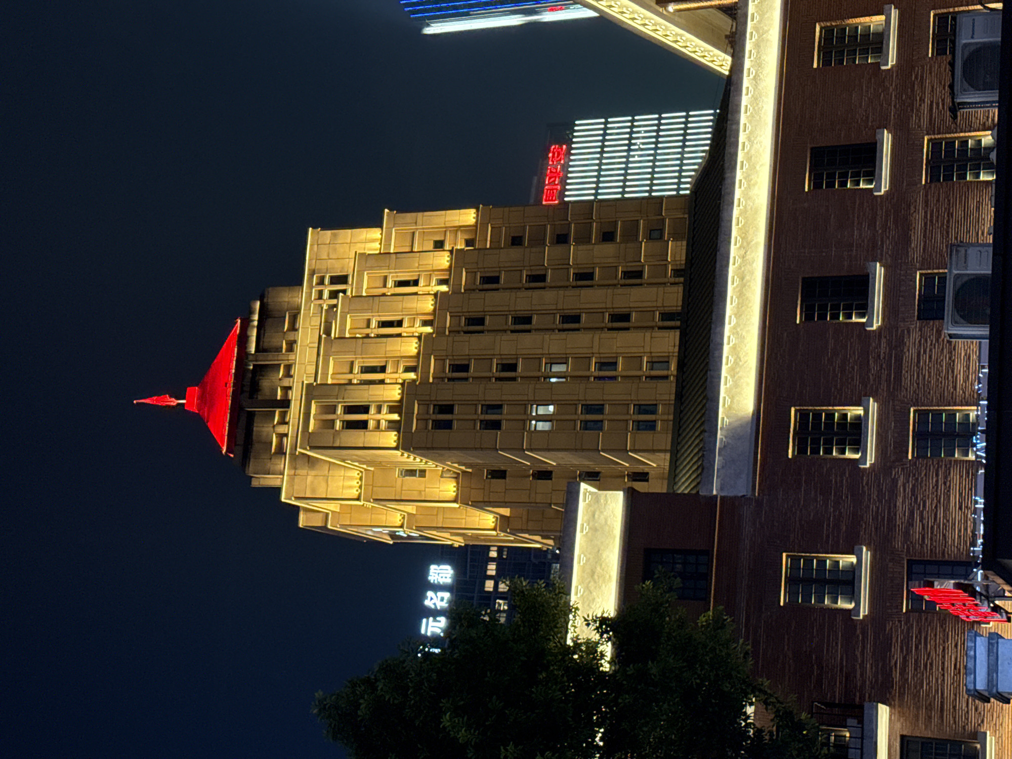 The image is a night-time, eye-level shot of a tall, illuminated building in Yuzhong District, China. The building features a distinctive red, pointed roof, topped with a red flag. The facade is golden-yellow, with many windows and vertical lighting strips. In front of the main building, a lower, brick building with illuminated windows is visible. A large tree with dark green foliage is on the left side of the frame, partially obscuring the view of the buildings. Several neon signs are visible in the background, one in white displaying Chinese characters and another in red with white Chinese characters. The sky is dark blue. There are no people or animals visible in the image.