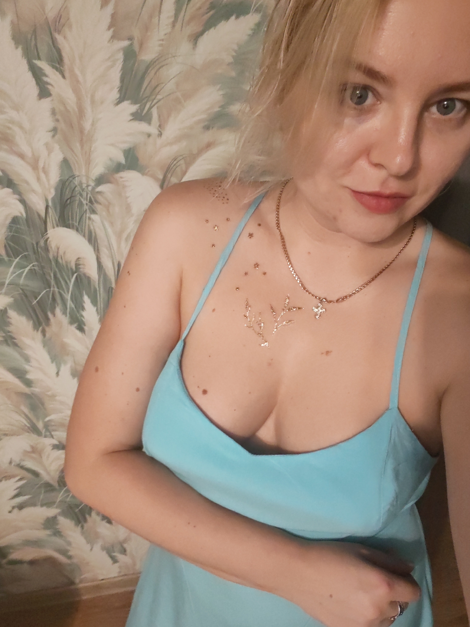 A woman with blonde hair and blue eyes is captured in a medium-close shot. She is wearing a light blue dress with thin straps and a delicate gold necklace with a cross pendant. Her chest is adorned with temporary gold glitter tattoos, one resembling a sprig and the other small stars. The woman is looking directly at the camera with a neutral expression.
The background features a wall covered in wallpaper with a botanical pattern of light-colored, feathery leaves against a muted green backdrop. The lighting appears to be indoor, casting soft shadows and highlighting the glitter on her skin. The overall scene suggests a casual indoor setting, potentially in a home. There is no visible text in the image. The context provided indicates the location is Izhevsk, Russia.