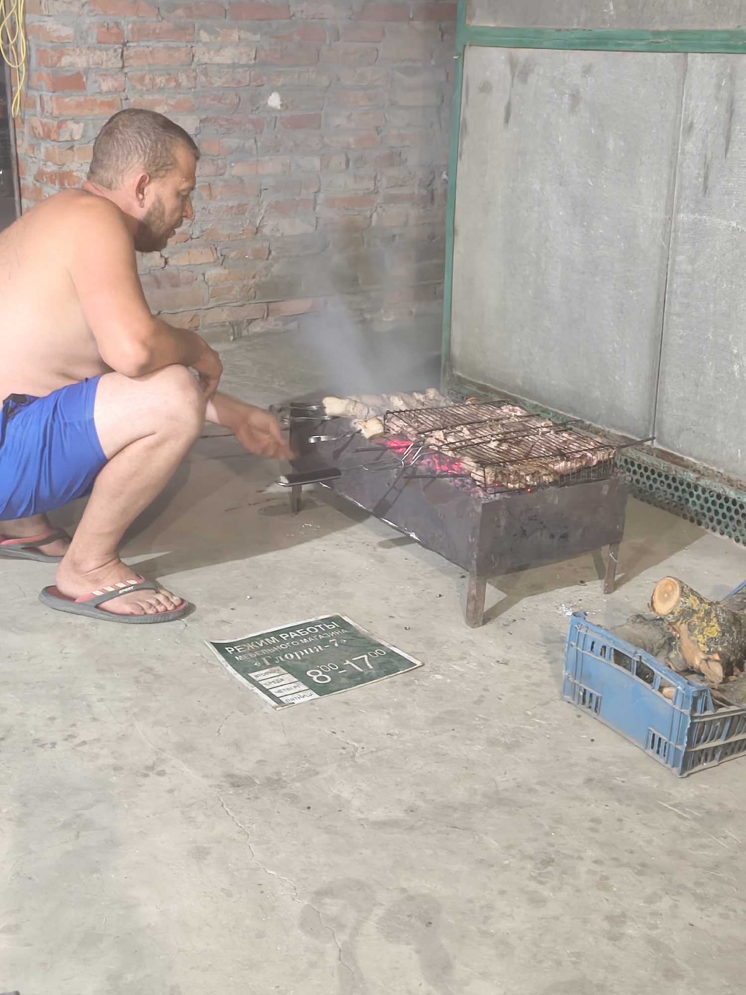 A man is squatting on a concrete floor next to a metal barbecue grill with meat cooking on it. The man is shirtless, wearing blue shorts and flip-flops. He is looking towards the grill. The grill is smoking and the coals are glowing red. In the background, there is a brick wall and a large metal structure with a green frame. On the ground, to the right of the grill, is a blue plastic crate filled with logs and a green sign with Russian text. The scene appears to be outdoors, possibly in a backyard or a communal outdoor cooking area in Novoaleksandrovsk, Russia.