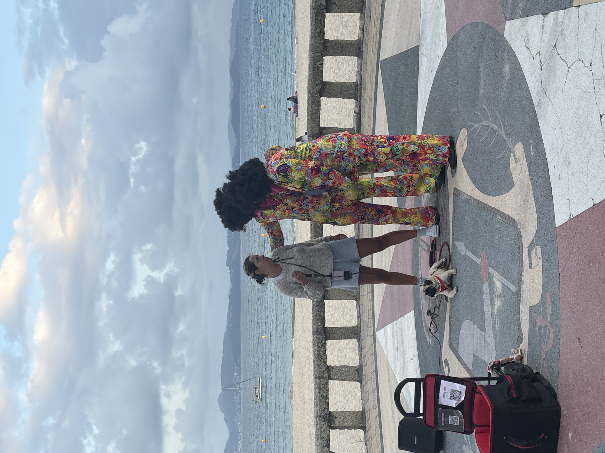 In Sanxenxo, Spain, on a paved promenade overlooking the sea, two individuals in matching vibrant, floral-patterned jumpsuits and large afro wigs stand with their backs to the camera. They appear to be interacting with a woman standing in front of them, holding a microphone. To the left of the woman, a small, light-colored pug dog is lying on the ground, leashed. In the foreground, a black and red wheeled cart with two QR codes displayed on it sits beside a black speaker. The background features a sandy beach, the ocean with a sailboat in the distance, and a cloudy sky.