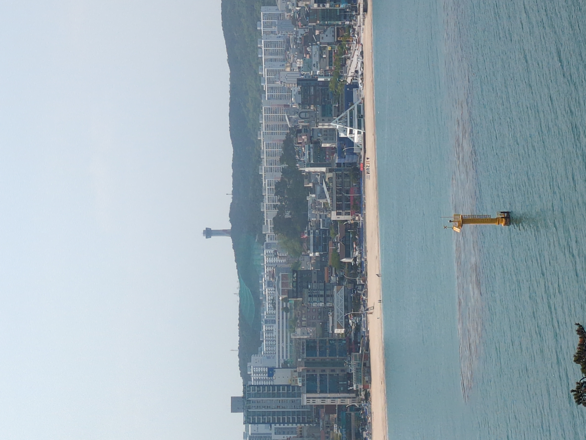 An aerial view looking east over the Haeundae district of Ulsan, South Korea. The coast with the beach and water runs vertically. Along the left side of the image is a line of dense, high-rise buildings that end at a tall, slender observation tower atop a mountainous ridge. The water is a blue-green hue. A yellow-colored structure sits in the water near the bottom.