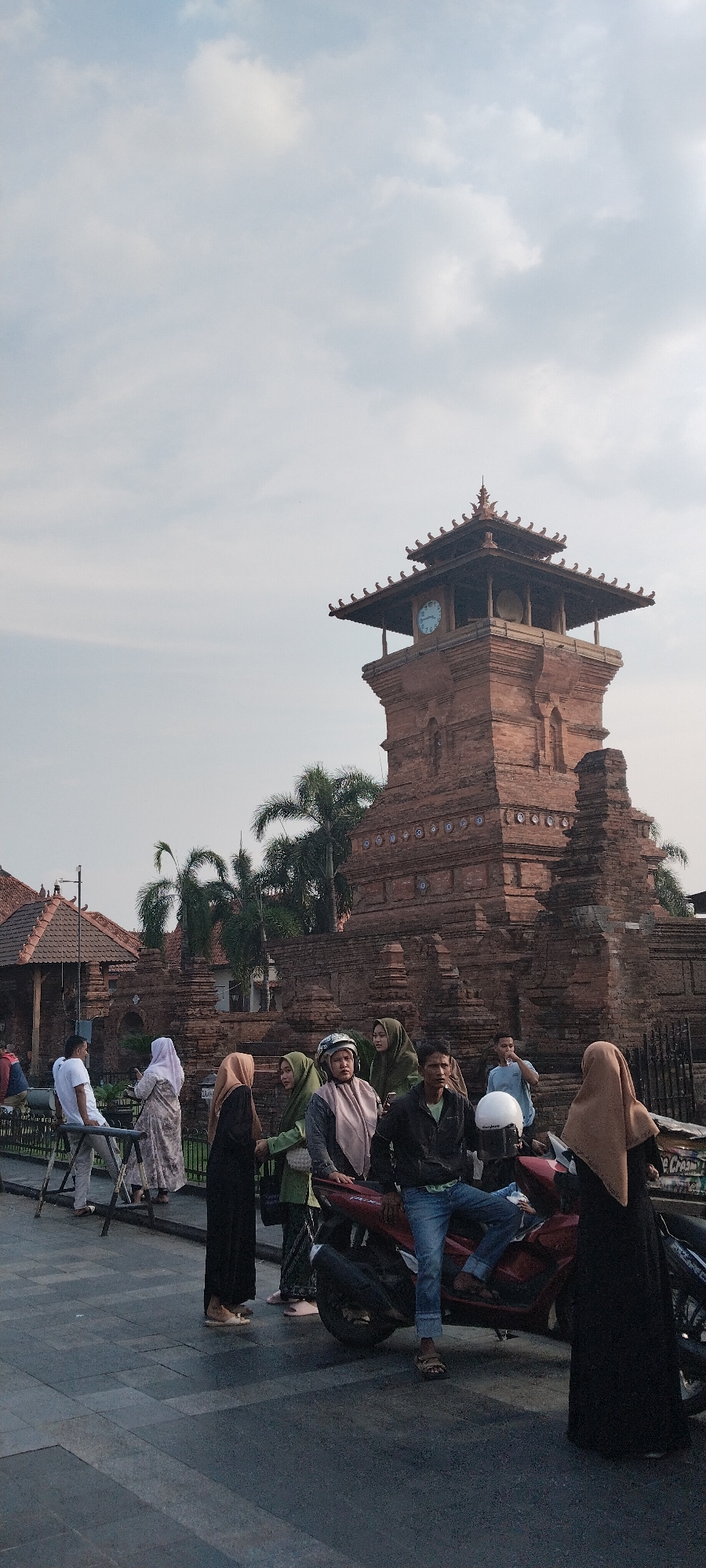The image captures a scene in the city of Kauman, Indonesia, featuring the iconic Menara Kudus Mosque complex. In the foreground, a group of people are gathered, some standing and others seated on a red motorcycle. The individuals are dressed in a mix of casual and traditional attire, with several women wearing hijabs. The focal point of the background is the striking Menara Kudus Mosque tower, constructed from red brick with a tiered, pagoda-like roof and a prominent clock face. Palm trees are visible behind the tower, adding to the tropical ambiance. The sky is overcast with soft clouds. The scene appears to be a public gathering area, possibly a tourist spot or a place of community interaction.