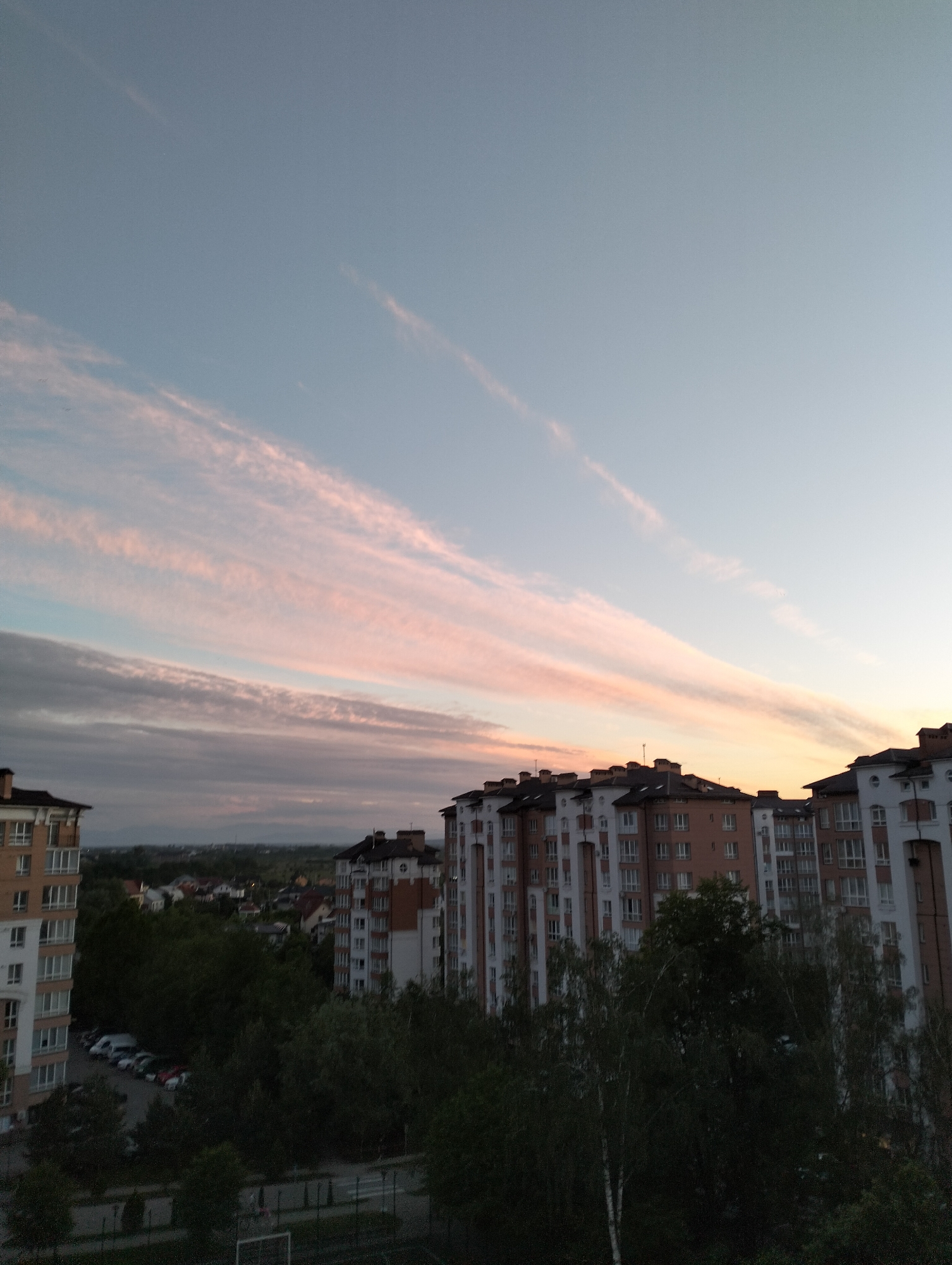 The image shows a view over Krykhivtsi, Ukraine. Multiple residential apartment buildings with white and orange facades are visible in the foreground. The buildings are partially obscured by trees. A parking lot with several cars is visible near the base of one of the buildings. A football field and a sidewalk can be seen. The sky is filled with long, pink and white clouds near the horizon, transitioning to a lighter blue color further up. The overall lighting suggests either sunrise or sunset.