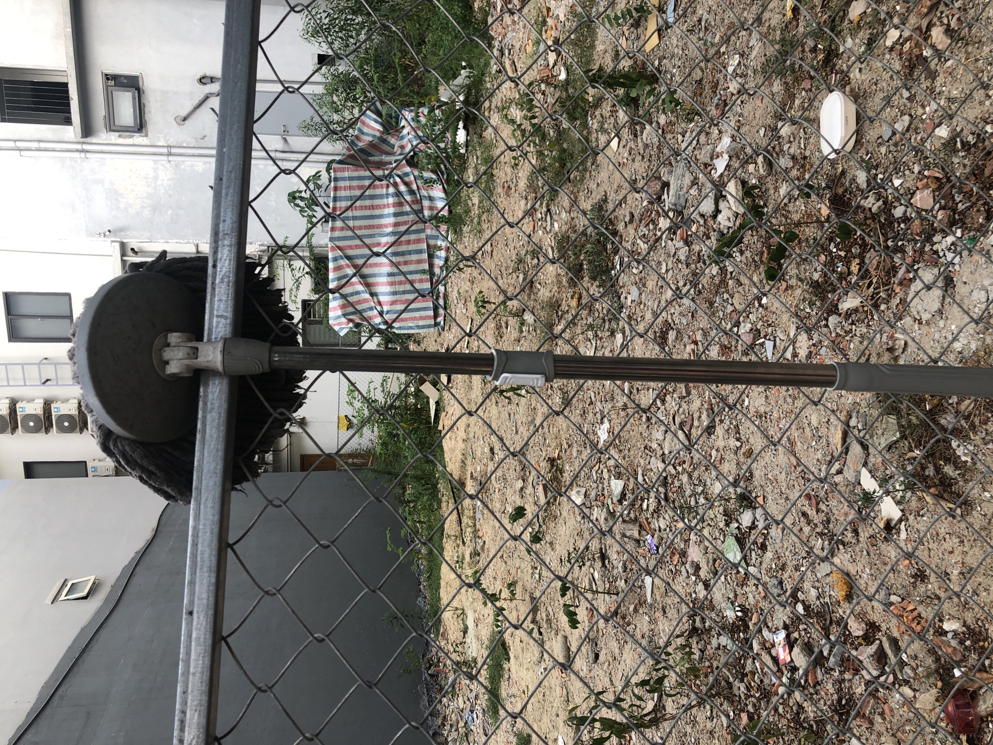 A mop with a dirty mop head is leaning against a chain-link fence in an urban area of Đà Nẵng, Vietnam. Behind the fence is a vacant lot filled with debris, including broken concrete, plastic, and discarded packaging. In the background, buildings can be seen, some with air conditioning units installed on their exterior walls. One building has a striped tarp covering something within the lot. The ground is mostly dirt and rubble, with sparse vegetation growing throughout. The scene appears to be a construction site or a neglected urban space.