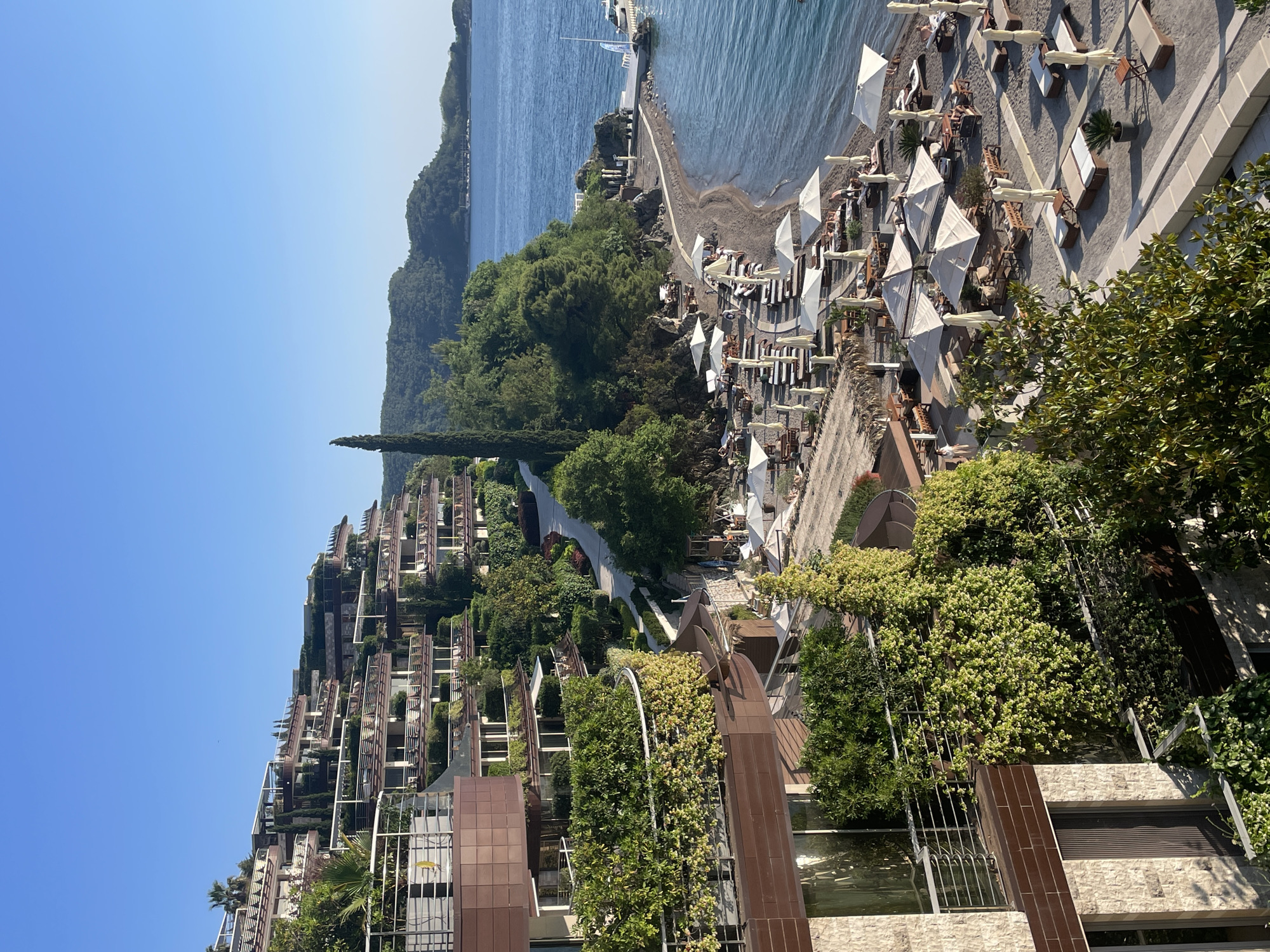 The image shows a coastal resort area in Budva, Montenegro. The scene is dominated by a terraced hillside with modern residential buildings. The architecture features extensive greenery. A pathway leads down to a beach area with numerous white sun umbrellas and lounge chairs. The Adriatic Sea stretches out to the horizon. A wooded peninsula extends into the sea. The sky is clear blue.
