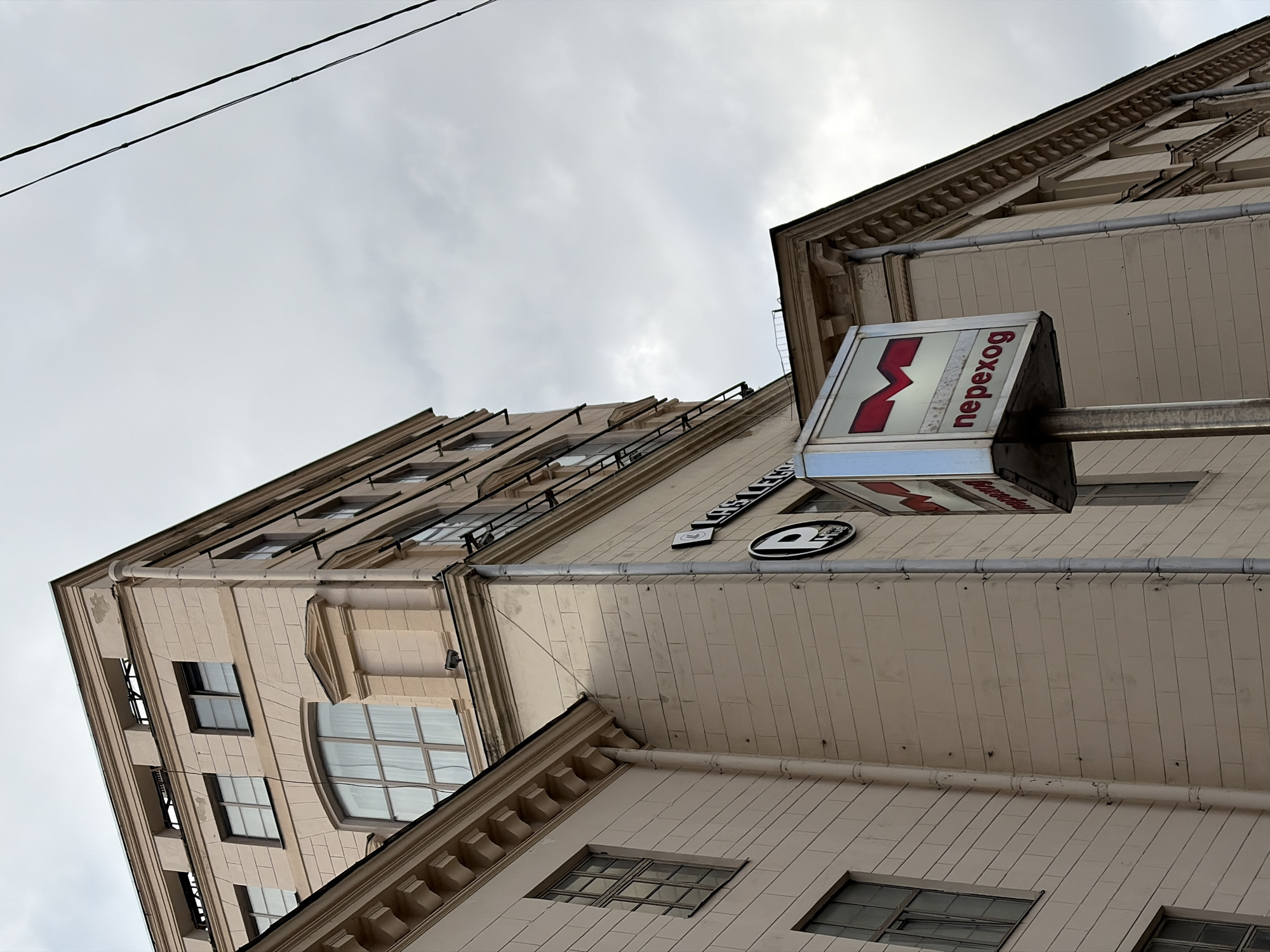 The media file shows a low-angle exterior view of a light-colored, multi-story building with ornate architectural details. The building appears to be made of stone or a similar material, with rows of windows and decorative cornices. Electrical wires run diagonally across the top of the frame against a cloudy, overcast sky.
A prominent sign with a red "M" logo and the word "переход" (perekhod, meaning "underpass" or "transition" in Russian) is mounted on the side of the building. Another sign, partially visible, displays Cyrillic text and a circular logo. The overall scene suggests an urban environment.
The time of day is indeterminate due to the overcast conditions, but it appears to be daytime. There are no people visible in the frame. The architecture and the Cyrillic text on the signs strongly suggest the location is in Minsk, Belarus, consistent with the provided location context. The weather is cloudy and appears to be cool. The mood is neutral, focusing on the architectural elements and signage of the building.
