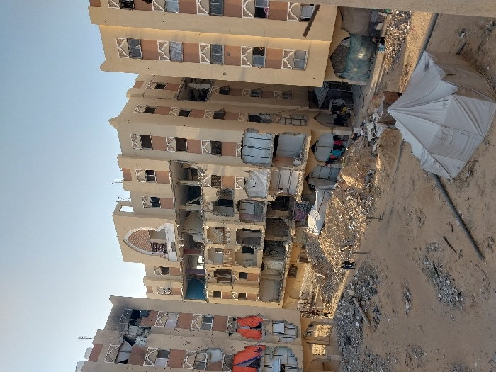 The image portrays an urban scene in Khan Yunis, Palestinian Territory, characterized by extensive structural damage to multiple multi-story buildings under a clear, light blue sky. Three large, beige-colored buildings, consistent with residential or commercial structures, are visible, all exhibiting significant damage. The central building is severely compromised, with multiple floors having their external walls blown out, exposing internal structures, and an arched facade element on its upper section is partially destroyed. The building to the left shows extensive damage, including blackened sections indicative of fire, numerous broken or missing window panes, some of which are covered with orange tarpaulins. The building to the right also shows signs of impact, with broken windows and exposed interiors.
The ground between these structures is unpaved, consisting of dirt and sand, heavily strewn with large quantities of construction debris, including concrete rubble, rebar, and miscellaneous shattered materials, forming substantial piles. In the foreground, a light-colored canvas tent, indicative of temporary shelter, is erected, with a large gray pipe running horizontally across the ground nearby. In the midground, two individuals stand near a large pile of debris. Further to the right, a solitary individual is seated near the base of the rightmost building. Near the base of the central building, colorful garments are hanging to dry, suggesting ongoing human presence. Green tarpaulins are also visible, creating makeshift enclosures near the lower levels of the buildings.