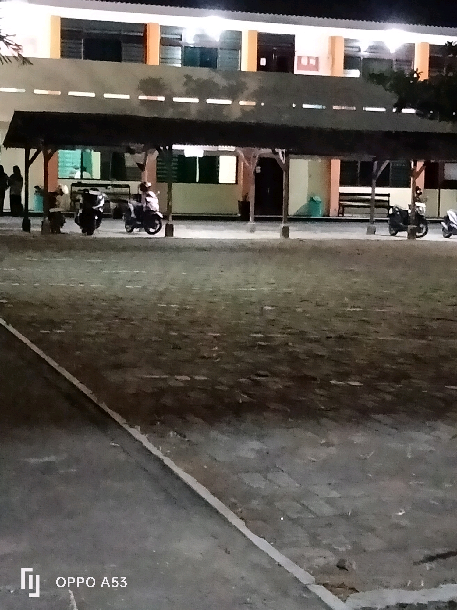 An outdoor, low-angle, night shot features a long, multi-story building with a covered walkway in front. The building has a pale beige facade with regularly spaced horizontal white lights along the top of the second story and orange vertical columns supporting the second story. The walkway has a dark, sloped roof supported by wooden posts. Several motorcycles are parked under the walkway. A few people are visible standing near the left edge of the frame, silhouetted against the lit interior of the building. The ground in front of the walkway appears to be paved with dark, textured material, possibly asphalt. A white line runs diagonally across the lower left of the frame. The bottom right corner of the image displays the text "OPPO A53". This scene is likely located in the city of Trenggalek, Indonesia, given the visible device branding which is common in that region.