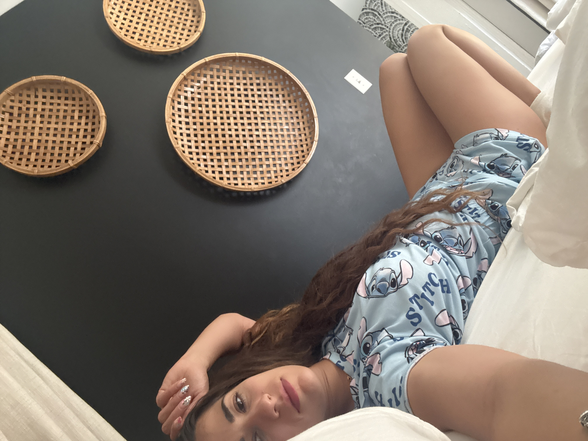 A young woman is lying on a white bed in a room with a dark wall decorated with three woven baskets. She is wearing light blue pajamas patterned with the character "Stitch" from the Disney movie "Lilo & Stitch." The woman has long, wavy brown hair and painted nails. The room appears to be indoors and well-lit. The location is not identifiable as Chicago, United States of America based on the available information.