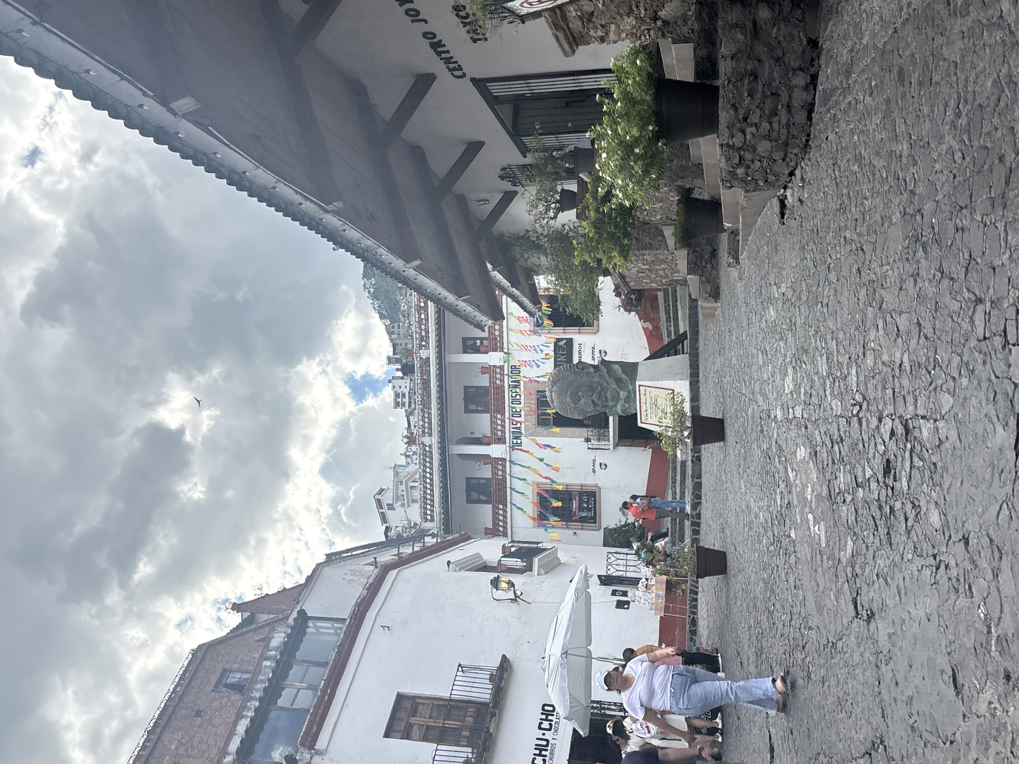 This is a full-shot, eye-level, outdoor photograph taken on a cobblestone street in Taxco, Mexico. The street is lined with white buildings, some with red trim and tile roofs. The sky is cloudy with a bird of prey flying in the upper left quadrant.
In the foreground, a woman in a white t-shirt and blue jeans is walking on the cobblestone street from left to right. She has light brown hair and is wearing a white cap. To her left, an open white umbrella covers a display of goods, likely for sale. Behind the umbrella, there are people browsing the merchandise.
Further into the scene, to the right of the woman, a large bronze bust statue is visible, mounted on a pedestal with a plaque. The bust appears to be of a man. Behind the statue, there is a staircase leading up to more buildings. Colorful festive banners hang across the street in front of one of the buildings.
To the right of the bust and staircase, a white building has a sign that reads "CENTRO JOY TAYCO". This building has a covered walkway with exposed wooden beams and decorative lights strung along the edge. Potted plants with green foliage and white flowers are placed on steps and along the walkway.
The architecture suggests a colonial or traditional Mexican style. The overall impression is of a bustling street scene in a historic town.