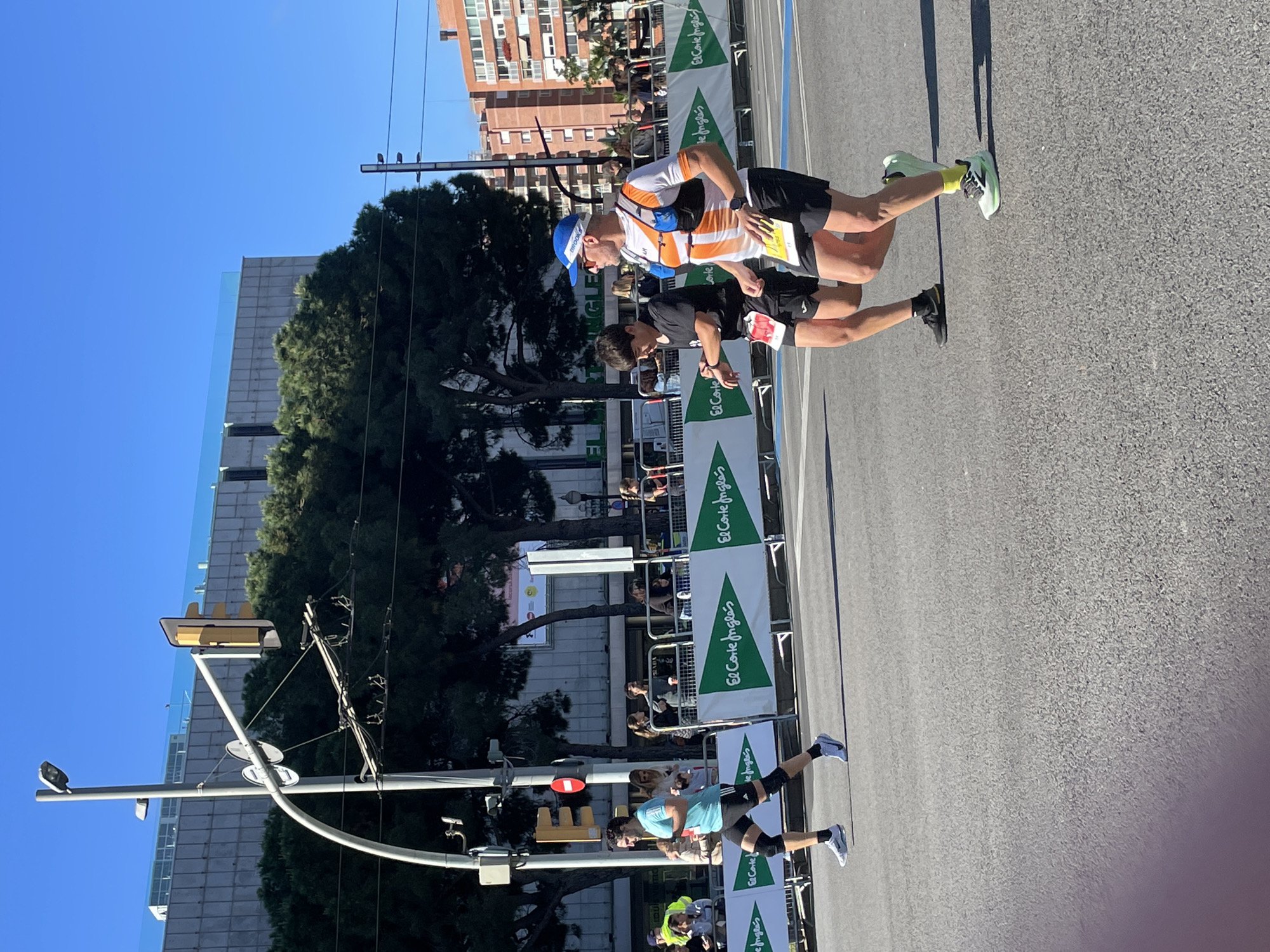 The image captures a road race or marathon taking place in an urban environment. In the foreground, runners are seen in motion on an asphalt road, their bodies angled with the exertion of running. A man in a white and orange striped shirt, blue cap, and hydration pack leads, closely followed by another runner in a dark shirt and shorts. The runners are wearing athletic shoes. Banners with the words "El Corte Inglés" line the side of the road, suggesting a sponsorship. Beyond the banners, spectators watch the race, their figures partially obscured by the barriers. A traffic light pole stands prominently in the background. A building with a glass facade can be seen. The sky is clear and blue, indicating a bright day. While the image does not have sufficient detail to confirm Barcelona, Spain, the presence of "El Corte Inglés", a Spanish department store chain, suggests a location within Spain.