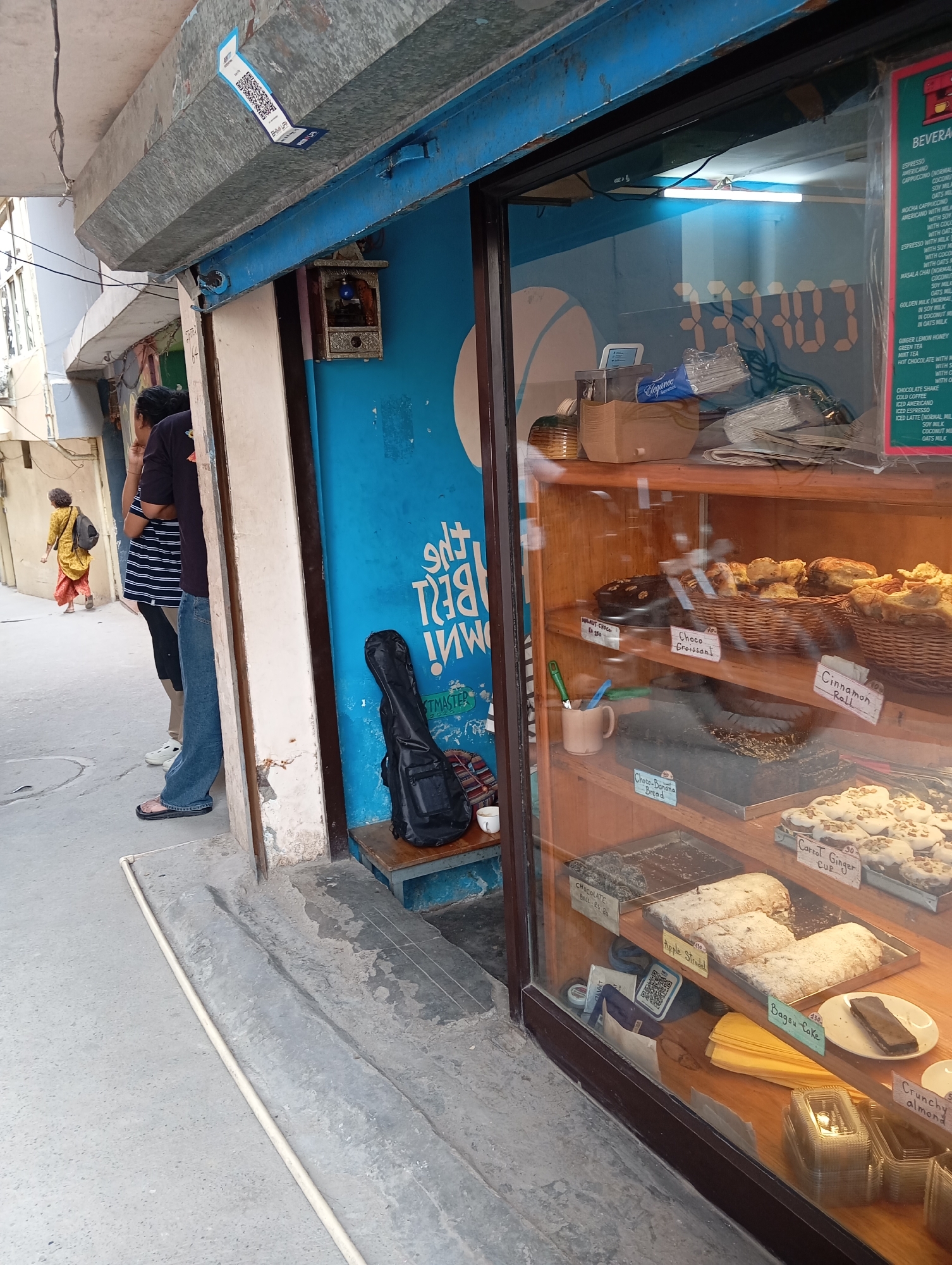 The image depicts a street-side bakery in what appears to be an older section of a city in India, as indicated by the style of dress of a woman walking in the background, and the visible QR code for local payment.
**Location:** Jonk, India (as specified)
**Setting:** A narrow street or alleyway.
**Environment:** The storefront is painted blue with white lettering. The adjacent buildings are older and constructed from concrete. There are concrete sidewalks and a street.
**People:** There are three people standing in the doorway, and one walking in the background.
**Subjects/Objects:** A bakery display case filled with various pastries (cinnamon rolls, chocolate croissants, carrot ginger cupcakes, etc.). There is a guitar case leaning against the blue wall next to the doorway, and a small shrine hanging above it.
**Actions:** Two people are standing in the doorway, seemingly talking. A woman is walking in the background.
**Interactions:** The people are likely customers of the bakery. The guitar case suggests a musician might be present or affiliated with the bakery.