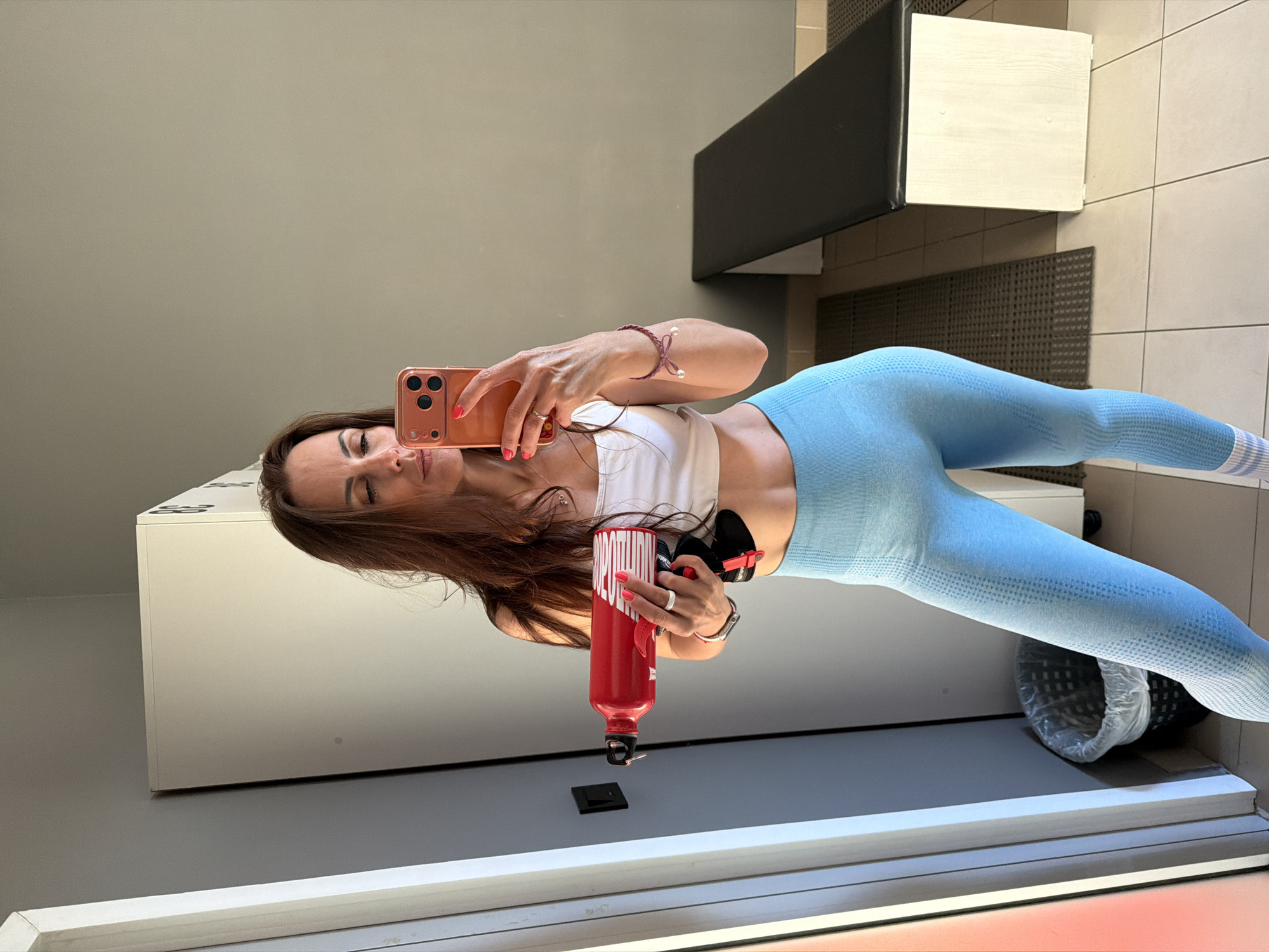 This image, taken in Birobidzhan, Russia, is a mirror selfie featuring a woman in what appears to be a gym changing room or fitness facility, rotated 90 degrees clockwise.
The subject is a woman with long, dark brown hair, looking into her coral-cased smartphone held in her right hand. Her fingernails are painted to match the phone case. She is wearing a white sports bra or crop top and light blue, textured seamless leggings, along with white socks featuring three blue stripes at the ankles.
Accessories include a silver watch with a red band and a delicate, dark pink braided bracelet with two white pearl-like beads on her left wrist. She wears a ring on her right ring finger and a white ring on her left ring finger. In her left hand, she holds a bright red water bottle with white text, possibly reading "SMOOTHIE."
The background indicates an indoor setting. There are light gray walls, a tiled floor with light beige squares and gray grout lines, and various pieces of furniture. These include white lockers or storage units, one showing partial text like "53" or "GB," and a black padded bench on a dark, textured mat. A black waste bin with a white liner is visible beneath the bench. A white trim runs along the bottom edge, likely part of the mirror's frame. The woman's expression is neutral, consistent with posing for a photograph. The scene suggests a post-workout or pre-workout moment in an athletic environment.