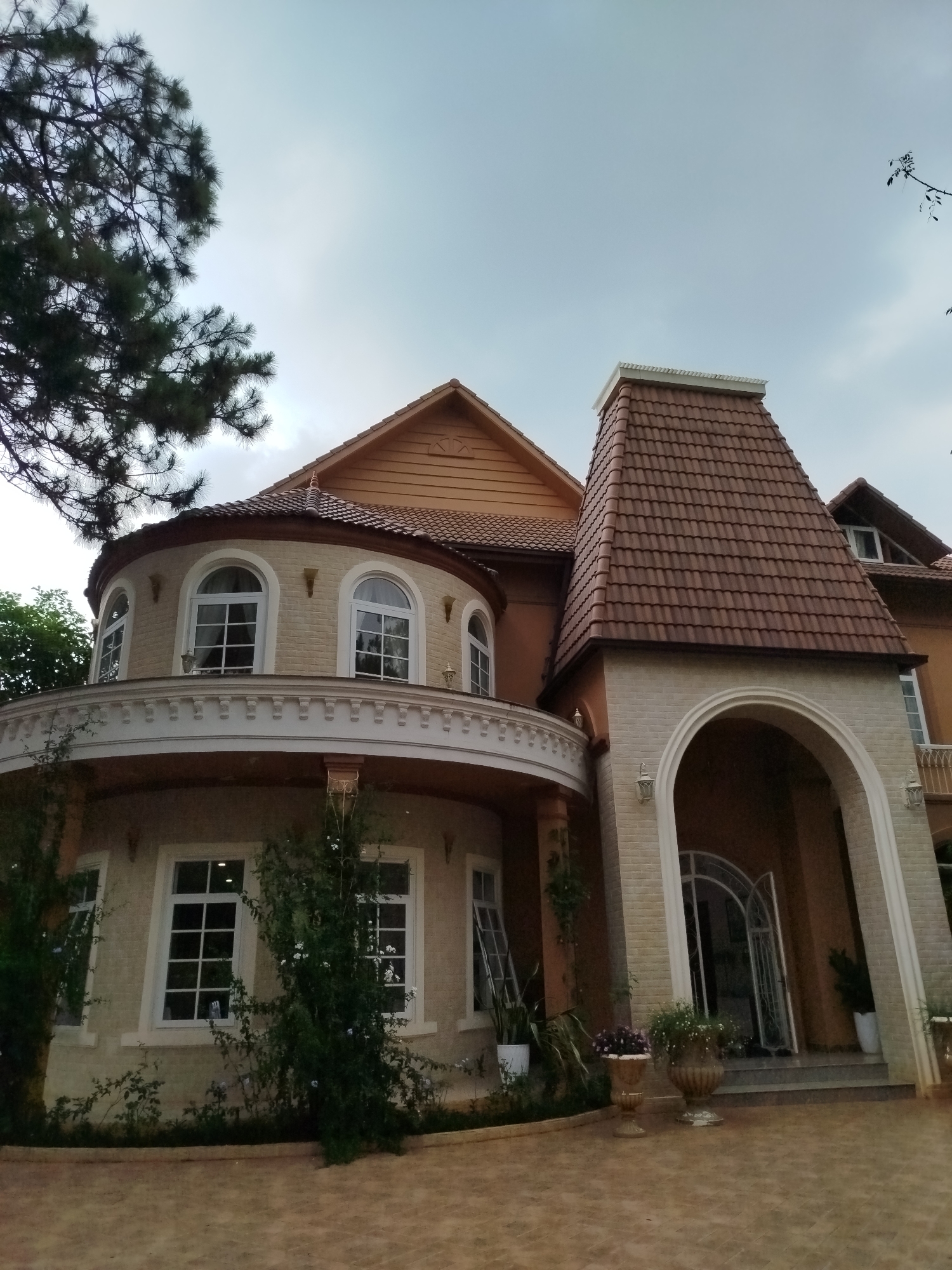 The image captures a distinctive two-story building in what is likely Da Lat, Vietnam, given the architectural style and foliage. The building features a rounded front section with arched windows on the upper level, and rectangular windows below. The facade is beige, with white trim around the windows. A brown tiled turret rises next to the rounded section, marking the entrance beneath a large archway. Plants are located in front of the building, adding to the picturesque scenery. The sky is overcast, and a large tree is on the left side of the frame.