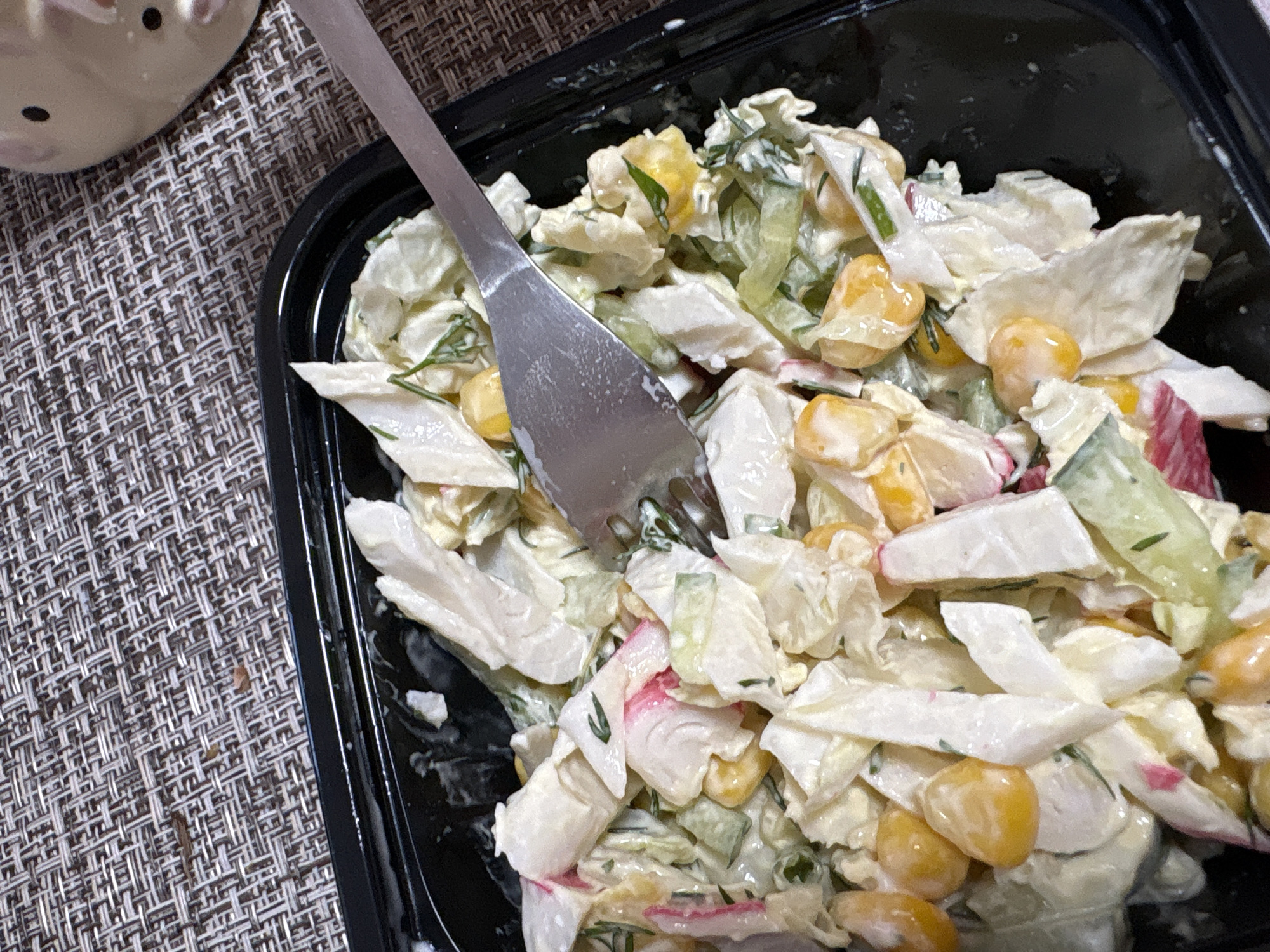 A close-up shot displays a black plastic container filled with salad. A silver fork is inserted into the salad. The salad consists of shredded cabbage, corn kernels, diced cucumber, and imitation crab meat mixed with a creamy dressing. The container sits on a textured, woven mat. A ceramic object is partially visible in the upper left corner. The city of Прикамский, Russia, is provided as the location, although it cannot be verified visually.