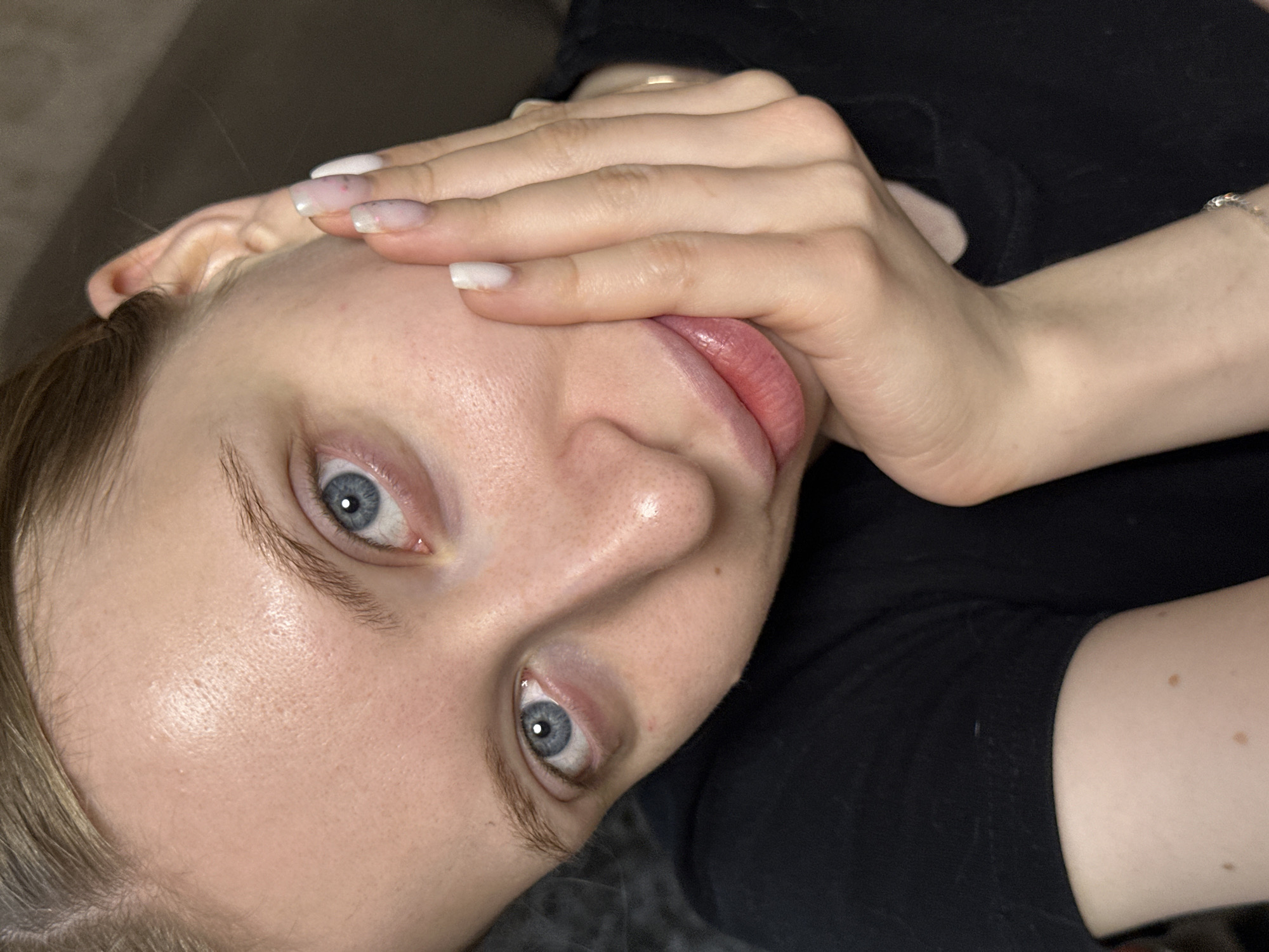 A close-up, angled selfie of a light-skinned woman with blue eyes. She is wearing a black shirt. Her right hand is partially covering her face, obscuring her forehead and cheek. The nails of that hand are manicured with a French tip style. A gold ring is visible on one finger of her right hand. A silver bracelet is visible on her right wrist. The background is blurred and dark. There is no information in the image to indicate it was taken in Chaikovsky, Russia.