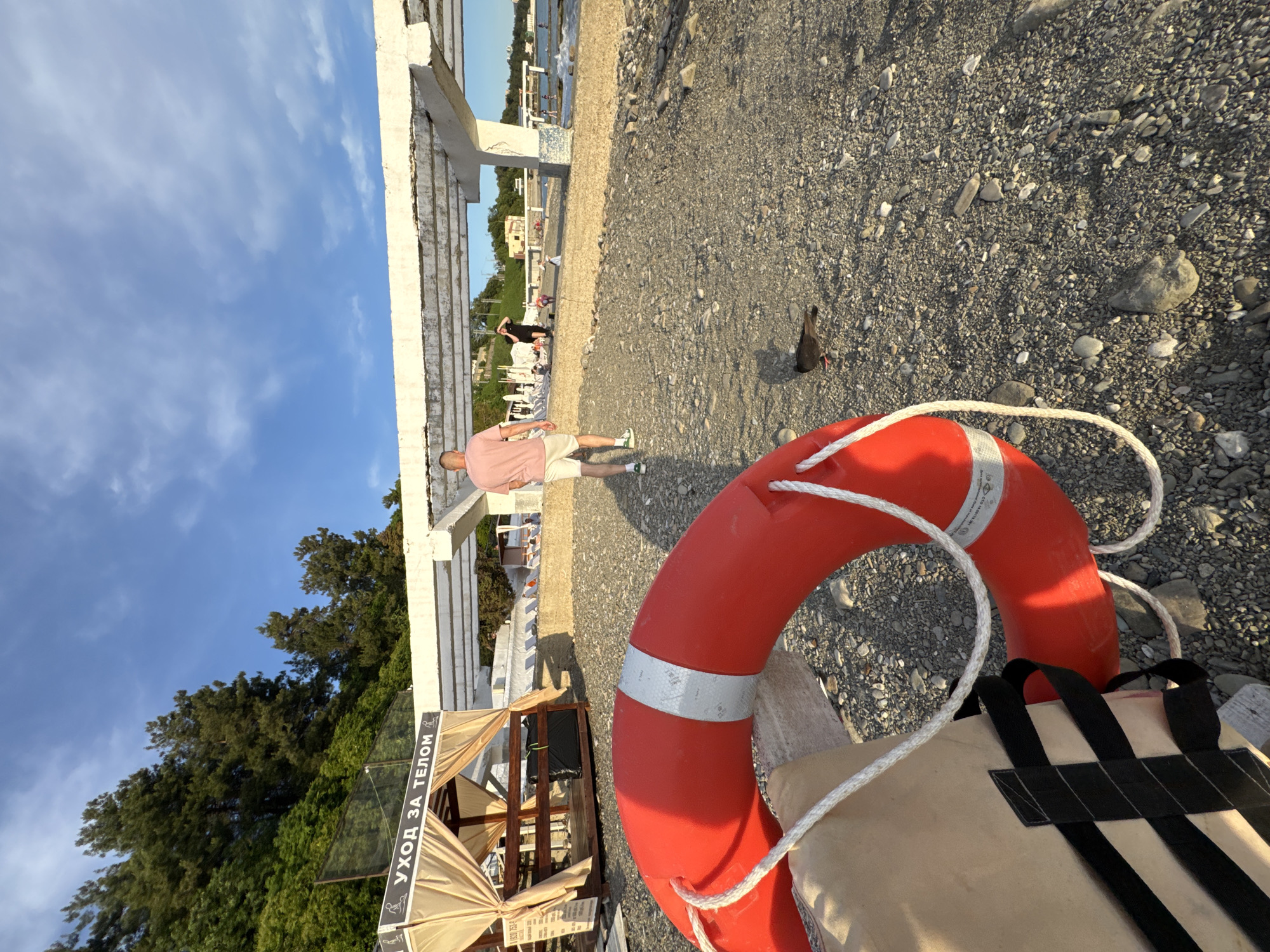 The image depicts a beach scene on a sunny day, likely in Sochi, Russia, judging by the Cyrillic text visible on a canopy that reads "УХОД ЗА ТЕЛОМ" (meaning "Body Care"). In the foreground, a red life preserver and a tan life vest are prominently placed on a pebbled beach. A small bird is seen foraging on the ground nearby.
In the midground, a man in a pink t-shirt and white shorts walks away from the camera. Further back, under a white concrete structure, several people are visible, some seated on beach chairs and others walking. The beach is lined with trees on a gentle slope. The sky is blue with scattered clouds. The overall impression is one of a relaxed, recreational environment by the sea.