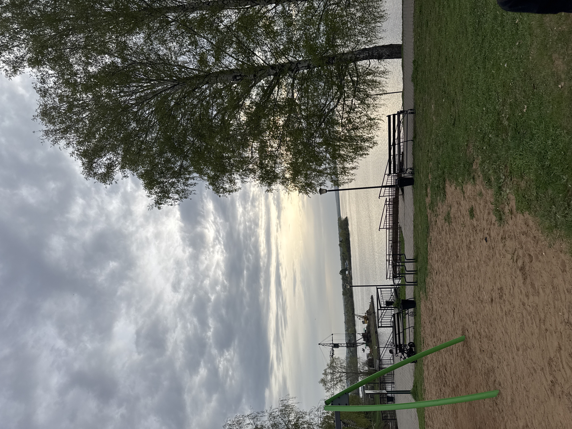 The image shows a view of the Kama River in the city of Osa, Russia, taken from a park or recreational area. The sky is overcast with dramatic clouds, partially obscuring the sun. A large birch tree with fresh green leaves dominates the upper part of the frame, its branches extending towards the river. The river stretches into the distance, with a thin strip of land visible on the opposite bank. Along the near bank, there is a waterfront area with black metal railings and benches. A green playground structure is in the foreground, situated on a sandy surface. There is a small lamppost in the middle ground. A patch of green grass is visible to the right. A piece of clothing is seen in the upper right corner. In the water, a crane can be seen by the waterfront. The general atmosphere is one of quiet tranquility under a cloudy sky.