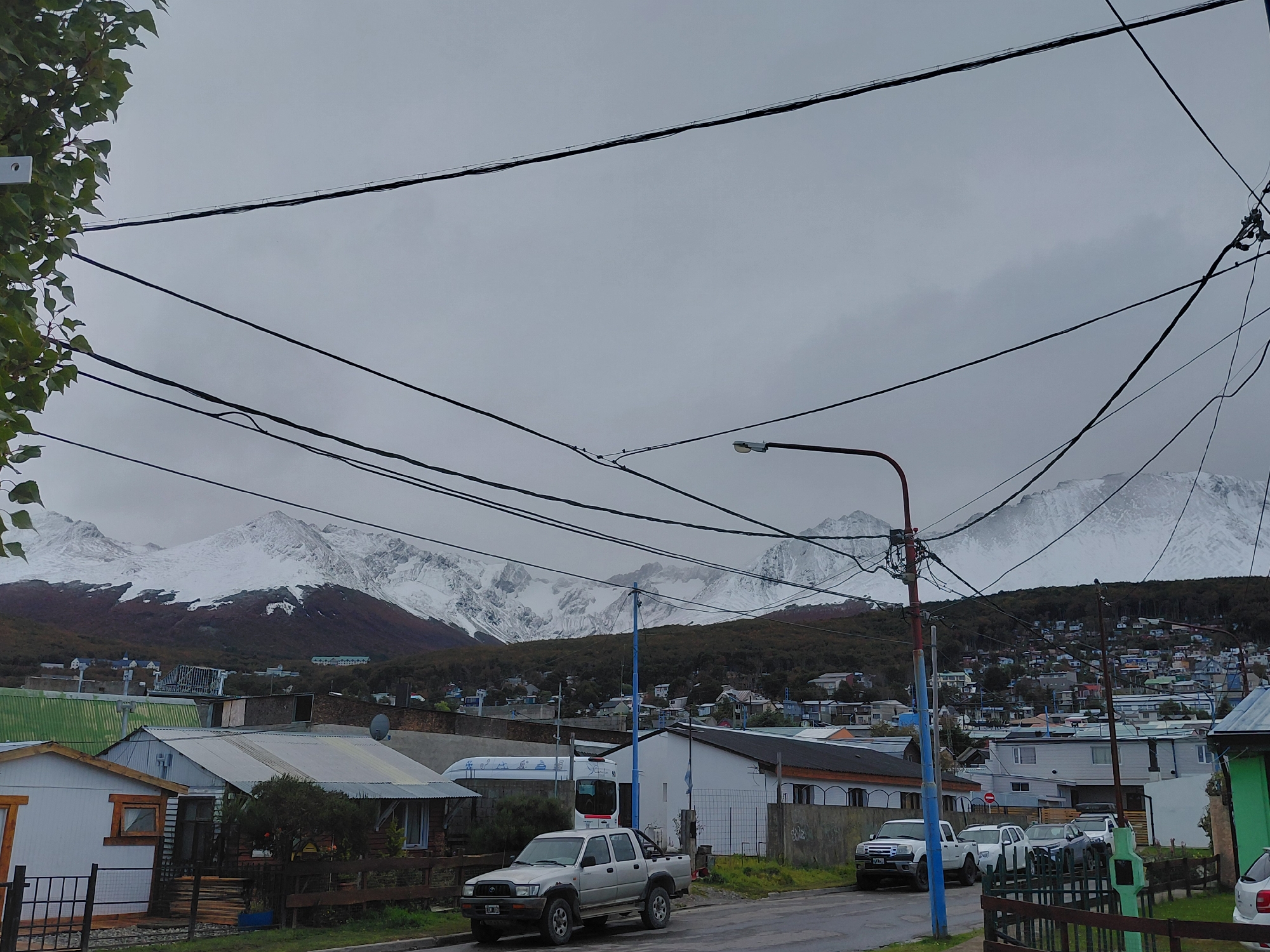 The image captures a street scene in Ushuaia, Argentina, under an overcast sky. In the foreground, a paved street is lined with a variety of single-story buildings, ranging from weathered shacks to more modern constructions. A silver pickup truck is parked on the street. Further back, a cluster of buildings spreads across a hillside leading to a line of snow-capped mountains on the horizon. Numerous power lines stretch across the sky. A lamp post with a mix of electrical and communications wiring stands prominently in the middle ground.