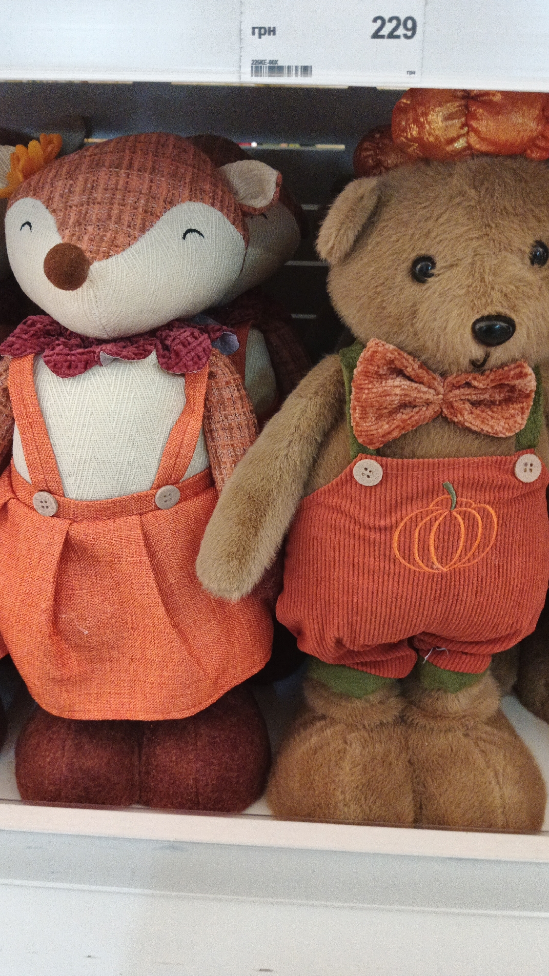Two stuffed toys are displayed side-by-side on a white shelf. The toy on the left is a fox with a textured orange and brown head and ears, and a white face with closed eyes and a small smile. It is wearing a cream-colored top with a ruffled burgundy collar, and orange overalls with two light gray buttons on the bib. The toy on the right is a brown teddy bear with a black nose and eyes, and a light brown bow tie. It is wearing a green undershirt and orange corduroy overalls adorned with an embroidered pumpkin design. Both toys have plump, stuffed feet. Above the toys, a white price tag is visible with the text "грн" and the number "229". The background is dark and out of focus. Based on the visible price tag indicating "грн", which is the abbreviation for the Ukrainian hryvnia, and the context of a store shelf display, this scene is likely located in a retail establishment within the city of Kryvyi Rih, Ukraine.
