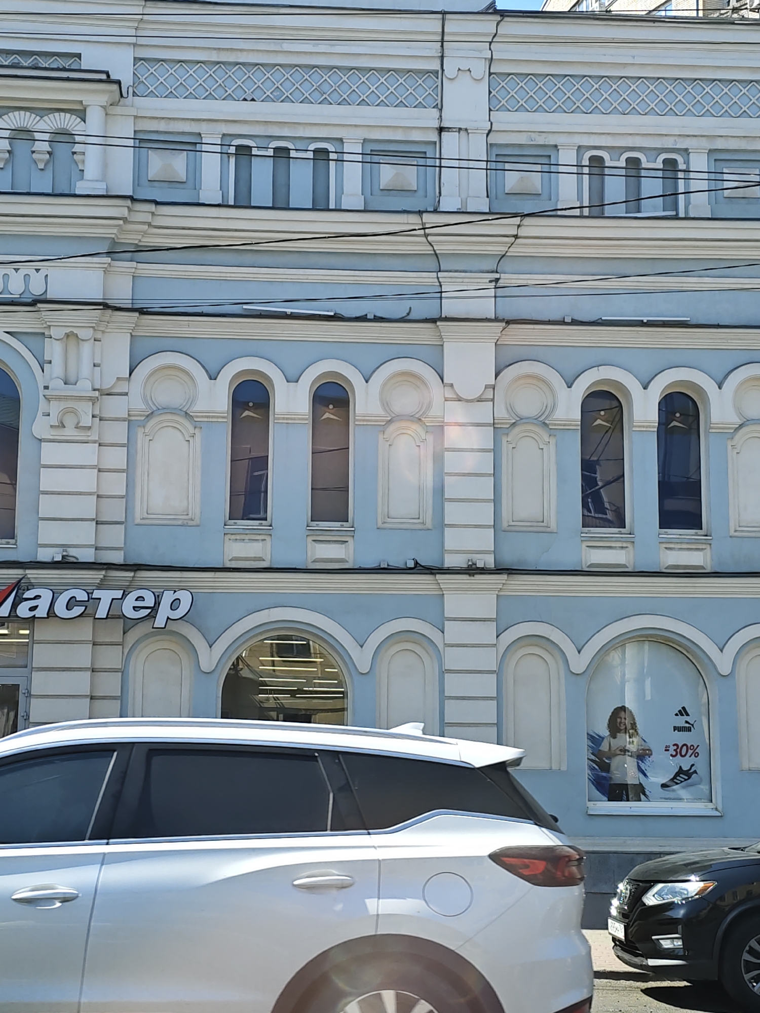 The image captures a section of a light blue and white building facade in Rostov-on-Don, Russia. The building exhibits ornate architectural details, including arched windows and decorative molding. A prominent sign with the white text "Мастер" is visible on the lower part of the building. In the foreground, the side of a white SUV is partially visible, positioned in front of the building. To the right, the front of a black Nissan SUV is also in view, parked on the street. A large shop window on the building displays an advertisement featuring a child, the Puma logo, and text indicating a "30%" sale. The overall scene appears to be a street view of a commercial establishment in a city.