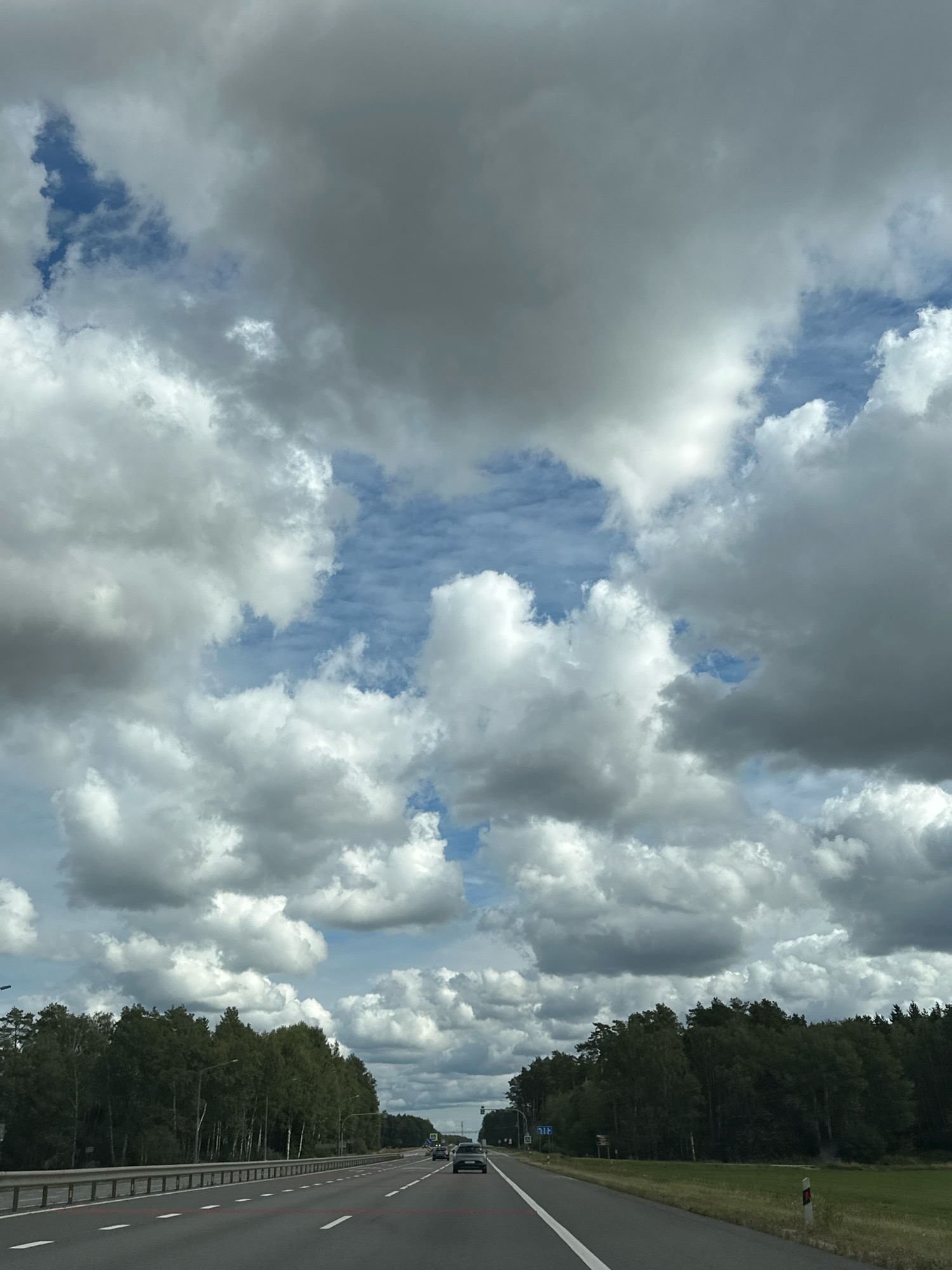 The image depicts a highway scene in the city of Пухавіцкі сельскі Савет, Belarus. A two-lane road stretches into the distance, flanked by a dense forest on both sides. The sky is filled with a dramatic display of clouds, ranging from bright white to dark grey, with patches of blue sky visible. A few cars are seen on the road, with one car prominently in the foreground on the right lane and another in the distance on the left lane. A guardrail runs along the left side of the road, separating it from the trees. Streetlights are also visible along the roadside. The overall impression is that of a typical day on a highway surrounded by nature.