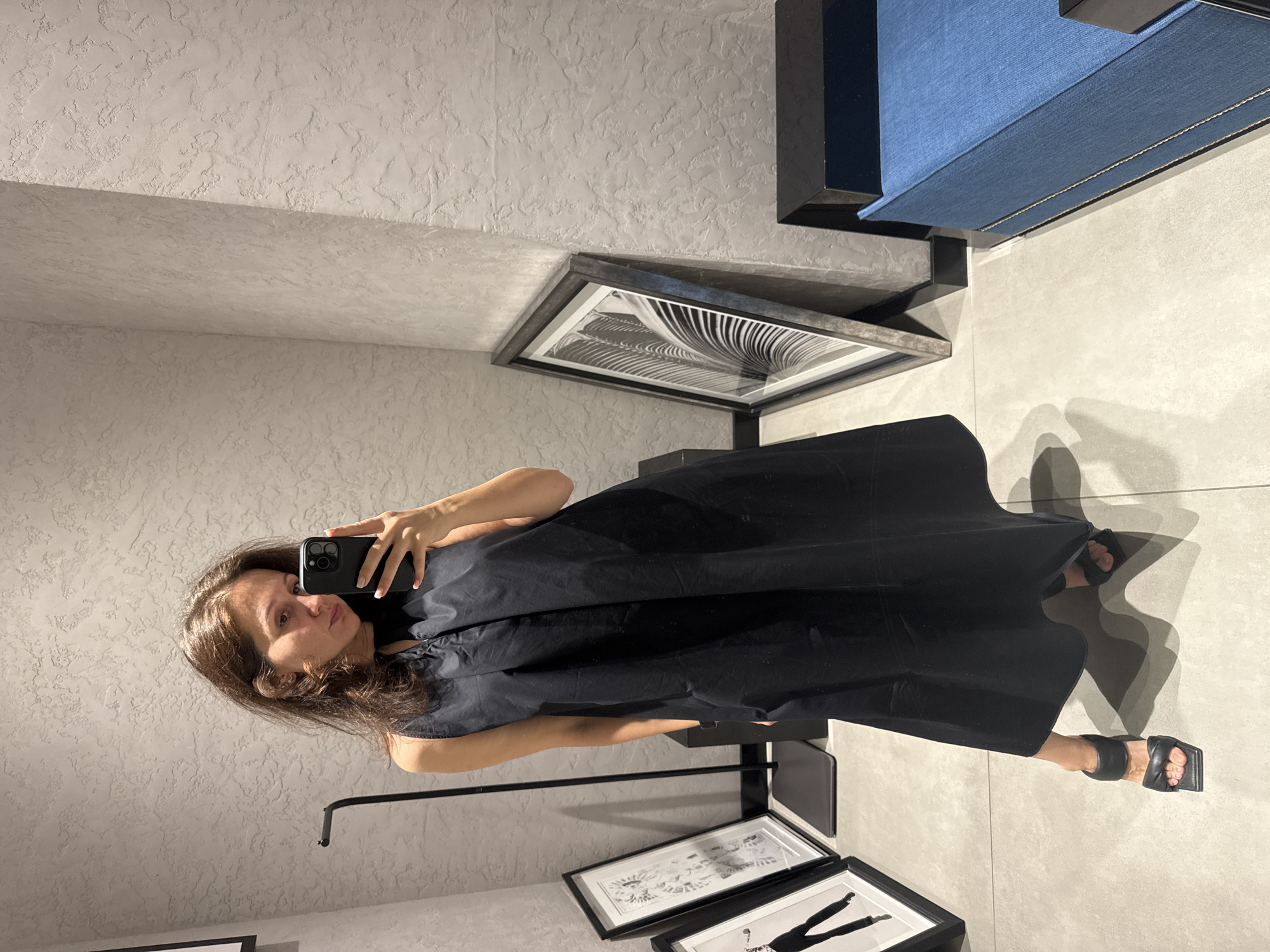 A woman is taking a selfie in what appears to be a retail space, likely a clothing store. She is holding a black smartphone and is wearing a loose-fitting black dress and black sandals. The store features textured walls, a modern art piece in a black frame, and display racks. The style of the retail space, including the art and furniture, suggests a high-end boutique. A blue upholstered bench is visible in the upper right corner. The image was likely taken in an upscale location and given the store's ambiance, Dubai, United Arab Emirates, is a plausible setting.