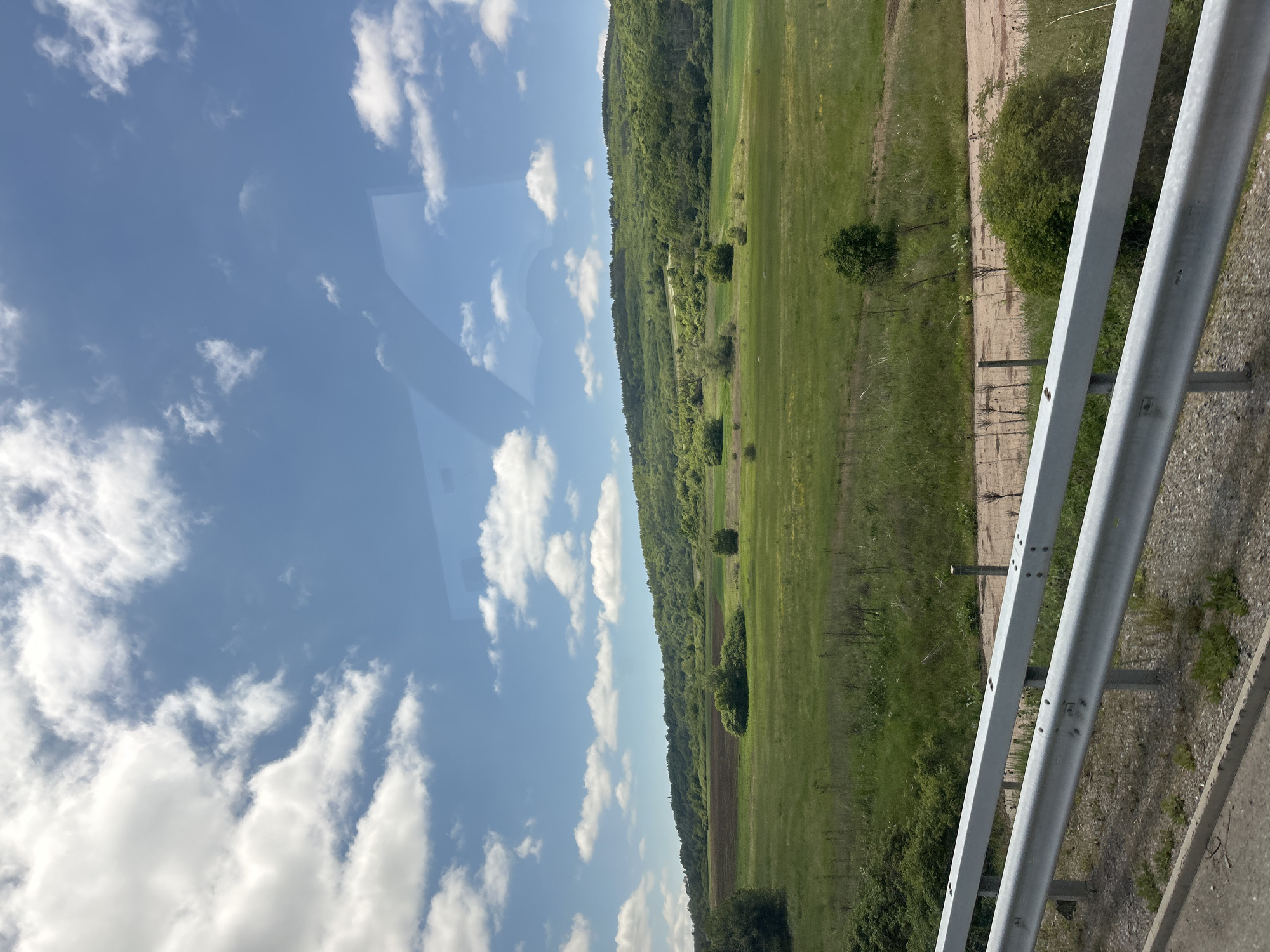 A view from a vehicle moving along a highway shows a landscape consisting of a green meadow extending to a tree-covered hillside. In the foreground, a metal guardrail indicates the proximity to the road. The sky above is blue with scattered white clouds. The location appears to be outside a city, in a rural area.