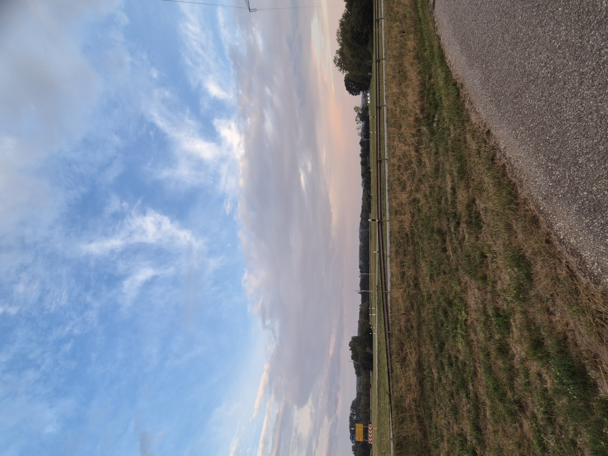 This is a photograph of a rural landscape, likely taken in Ergolding, Germany, at dusk. The sky is a dominant feature, filled with a mix of blue sky and wispy white and pink clouds, suggesting the sun is setting or has recently set. Below the sky, a flat, open field stretches out, with dry, golden-brown grass in the foreground and greener patches further back. A dark line, possibly a road or railway track, runs horizontally across the midground. Beyond this, a line of trees forms a dark silhouette against the horizon. A yellow directional sign with black lettering is visible on the left side of the image. In the far right, a tall, metallic structure, likely a power pylon, stands against the sky. The foreground of the image shows a rough, grey, gravelly surface, possibly a path or shoulder of a road, angled from the bottom right corner upwards. There are no people or animals visible in the scene. The overall impression is one of a quiet, natural setting during twilight.