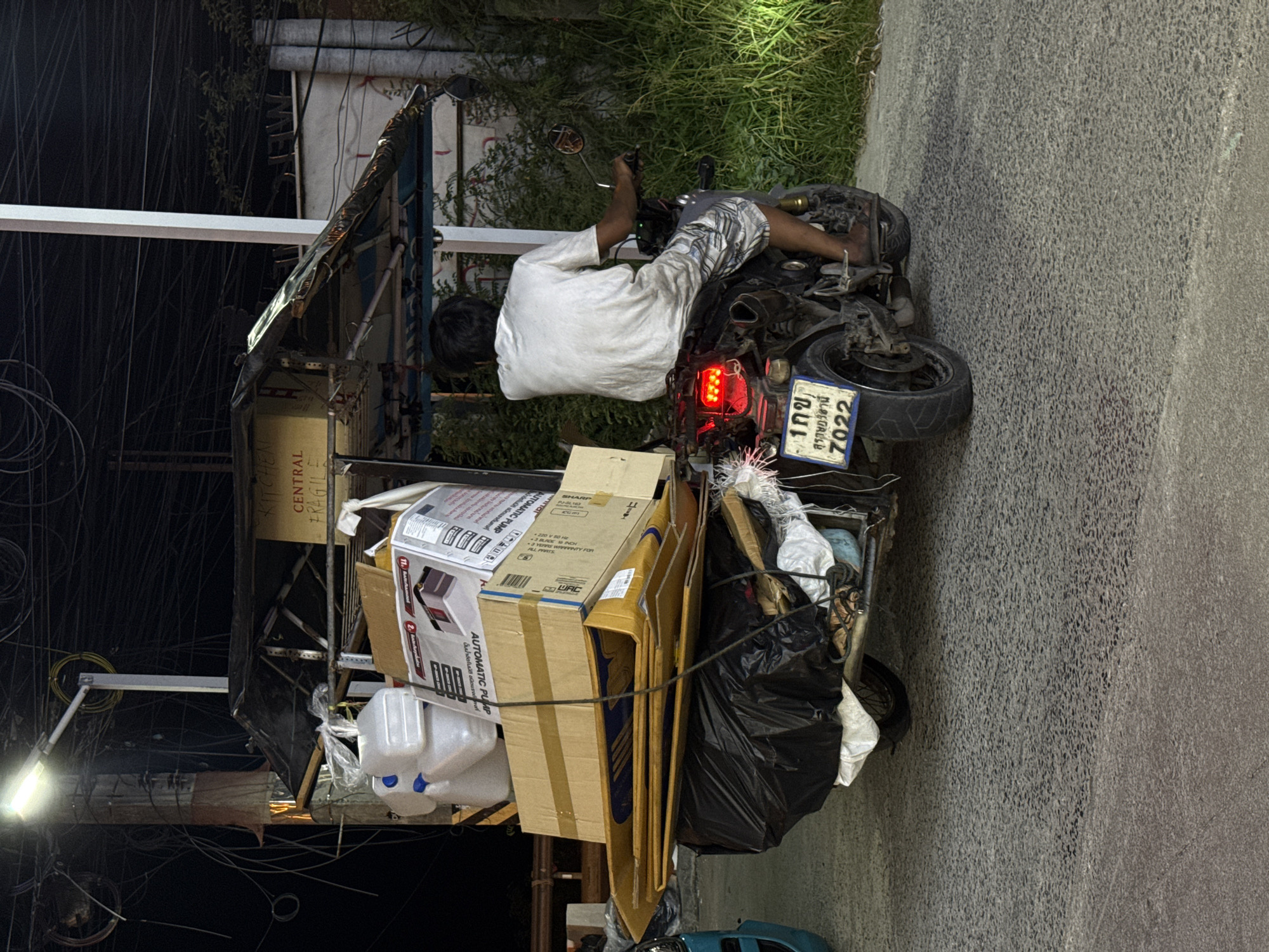 A nighttime, medium shot captures a modified motorcycle with an extended cargo platform, laden with various items, traveling on a paved street in Baan Bo Phut, Thailand. A person wearing a white t-shirt and patterned shorts is seated on the motorcycle, facing away from the camera, operating the vehicle. The cargo includes several white plastic water containers stacked on top of cardboard boxes, some of which are labeled. A black tarp covers some of the lower cargo. The motorcycle has a license plate with Thai characters and the number "7022". The background features a street lamp illuminating a dense network of overhead electrical wires, a building with a corrugated metal roof structure over the cargo, and overgrown vegetation along the right side of the street. The asphalt street surface is visible in the foreground.
