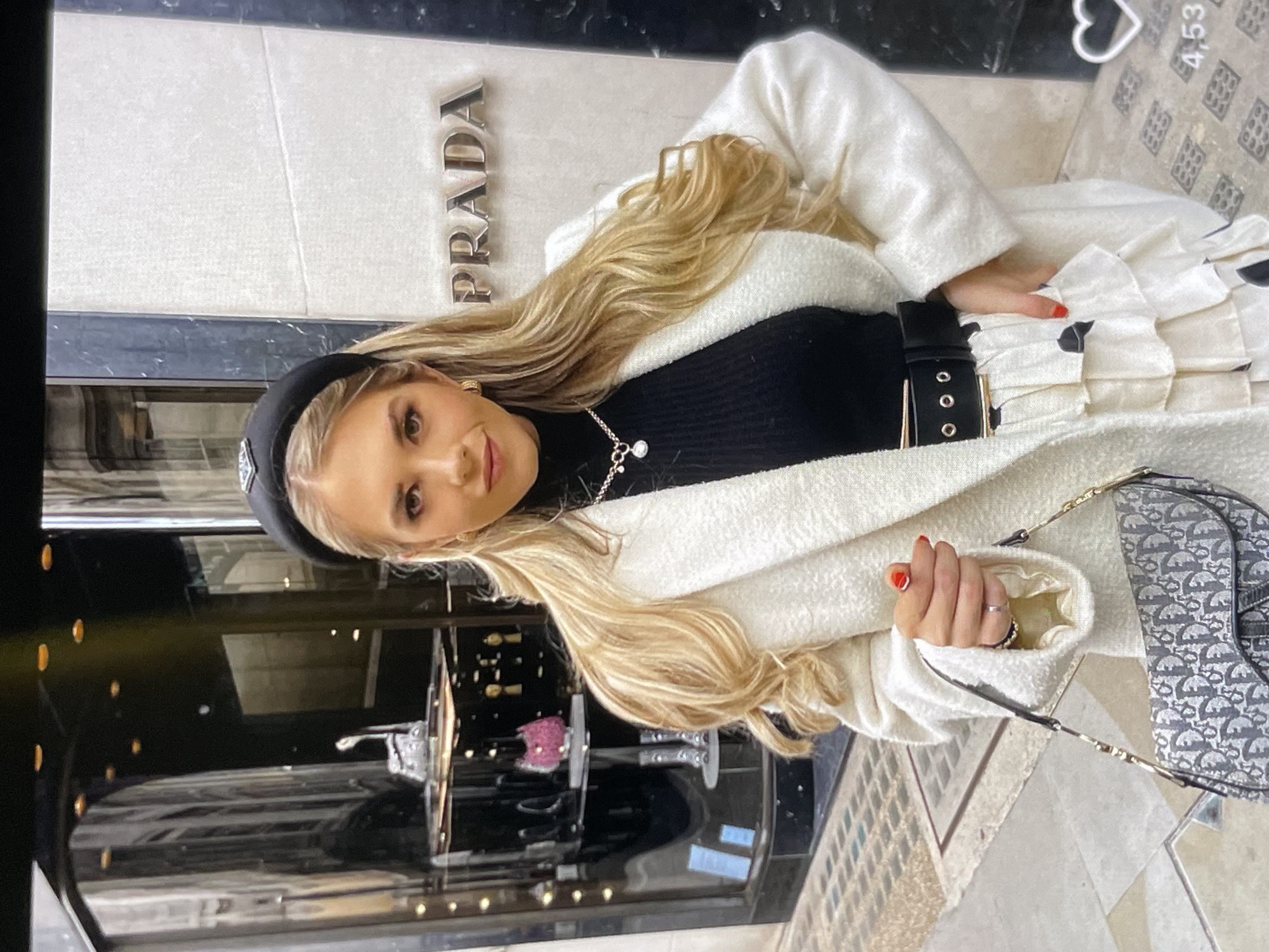 A blonde woman stands in front of a Prada store. She is wearing a white textured coat, a black turtleneck, a black headband with a Prada logo, a statement necklace, a black belt with a gold buckle, and a white tiered skirt. She is holding a Dior saddle bag. The background shows the storefront window display with mannequins and merchandise, and the Prada logo on the building. The ground is paved with cobblestones.