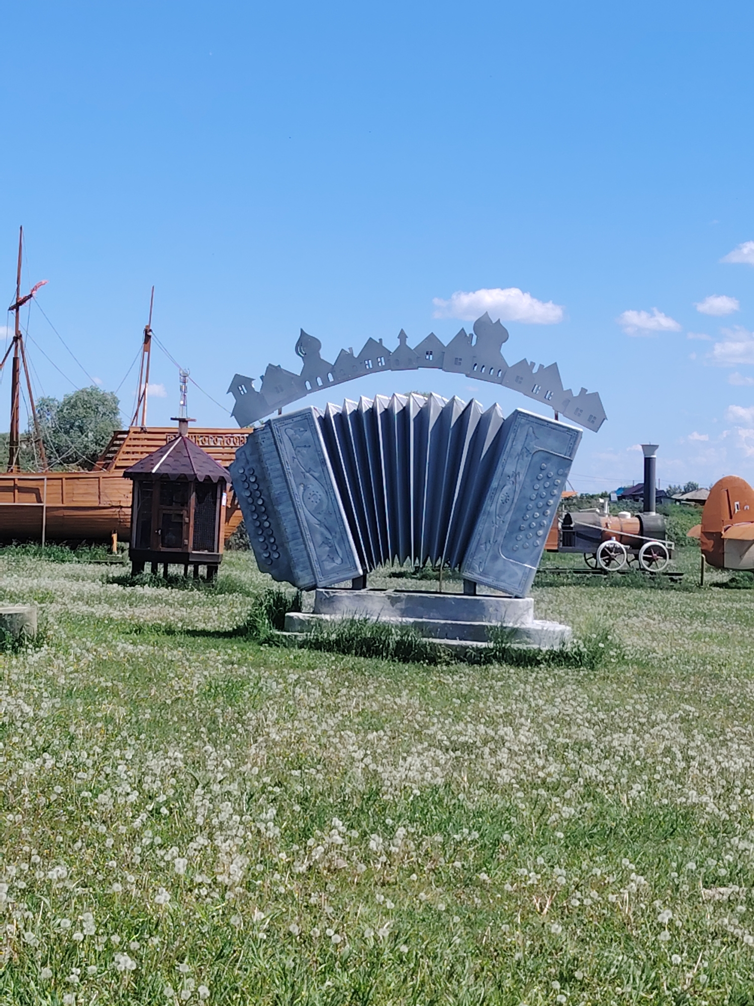 The image depicts an outdoor scene with a large, stylized metal sculpture of an accordion in the foreground, set against a backdrop of a blue sky with scattered clouds. The accordion sculpture features a decorative cityscape along its top edge. To the left of the accordion, there is a wooden boat with masts, and a small wooden structure with a peaked roof. Further in the background, a vintage-looking train and an orange, rounded object are visible, along with some distant buildings and trees. The ground is covered in green grass with numerous white dandelion seed heads, suggesting a meadow or park-like setting. The overall impression is of an open, rural or semi-rural environment.
