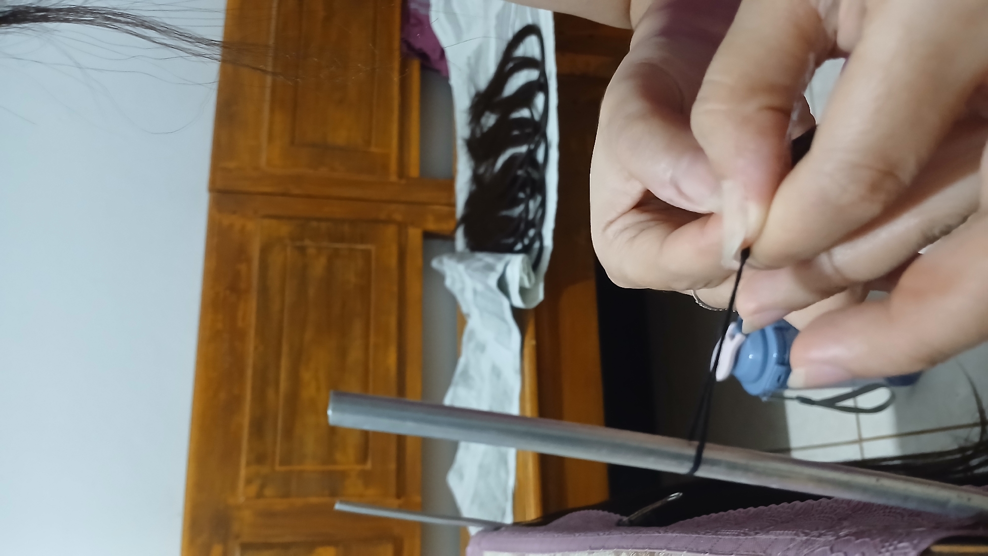 In Sampang, Indonesia, this image captures a close-up view of a person's hands as they tie a black string around a silver pole. The background reveals a wooden cabinet with two drawers slightly ajar, upon which a white cloth is laid out. Several bundles of dark hair are arranged on the cloth. A light blue water bottle with a gray strap is visible behind the pole. The lighting suggests an indoor setting.