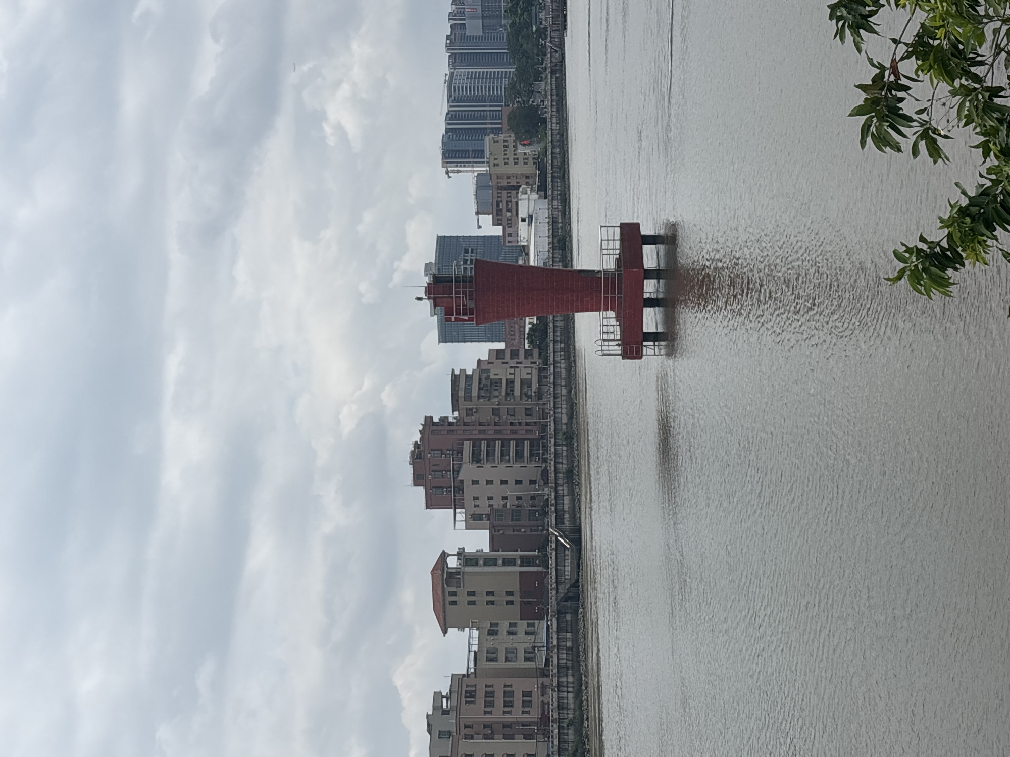 In Nanhai District, China, a red lighthouse stands in the middle of a body of water. The lighthouse is conical in shape with a white railing around its top. It is supported by a square base that rests on pillars submerged in the water. In the background, a row of buildings forms a cityscape along the waterfront. The sky is overcast with clouds. The water is calm, with small ripples on its surface. There are no people visible in the image.