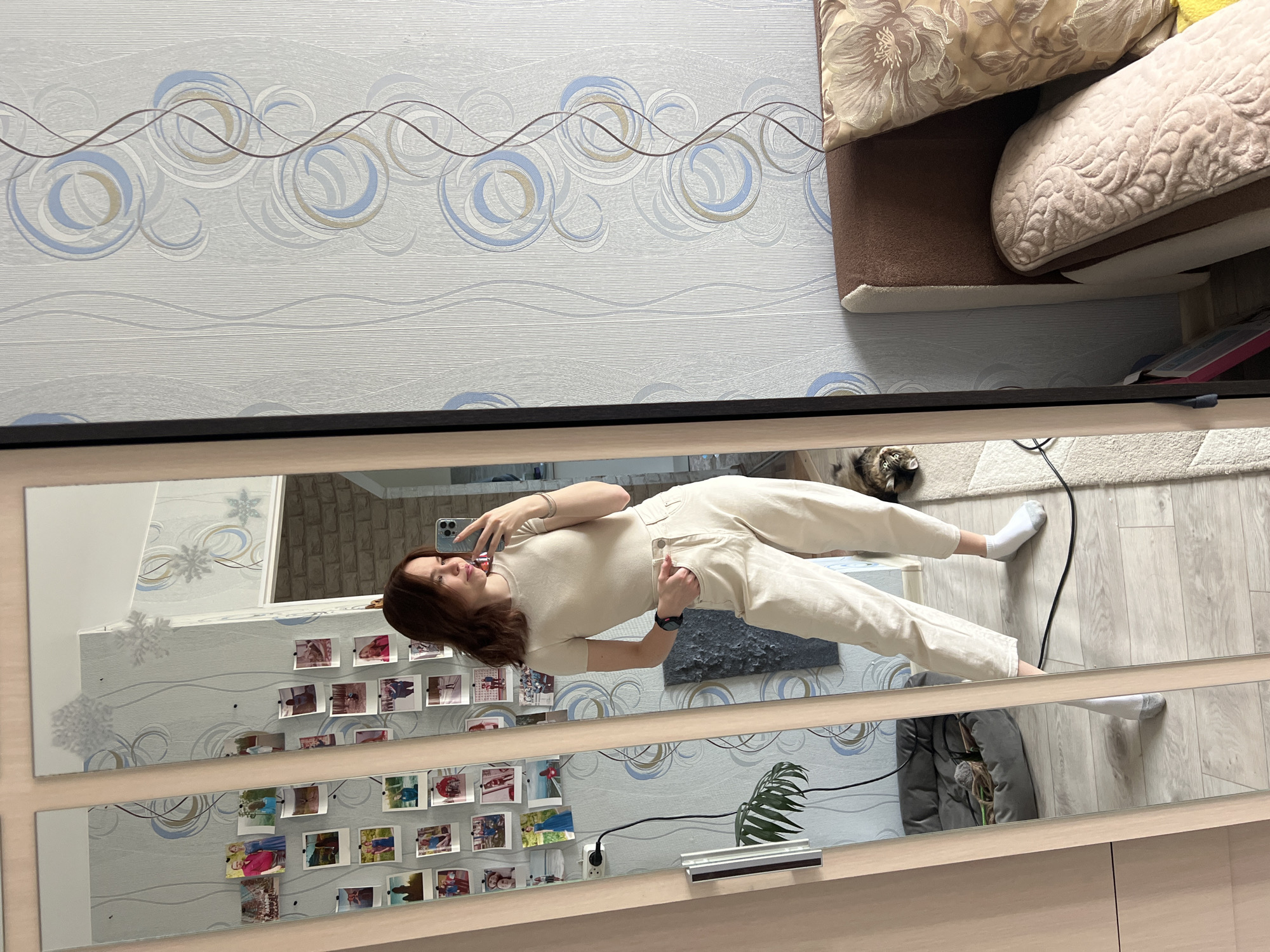 A full-length mirror reflects a young woman in a pale-colored, short-sleeved shirt and trousers taking a selfie with a phone. Her hair is brown. She is standing on a light wood floor with a white rug nearby. A cat is visible in the background. The mirror is mounted on a wall covered with patterned wallpaper. Small photographs are affixed to the mirror's frame. A plant is near the bottom of the frame. The reflections imply the photo was taken indoors in an apartment in Chaikovsky, Russia. Pillows are piled on a couch in the upper right of the frame.