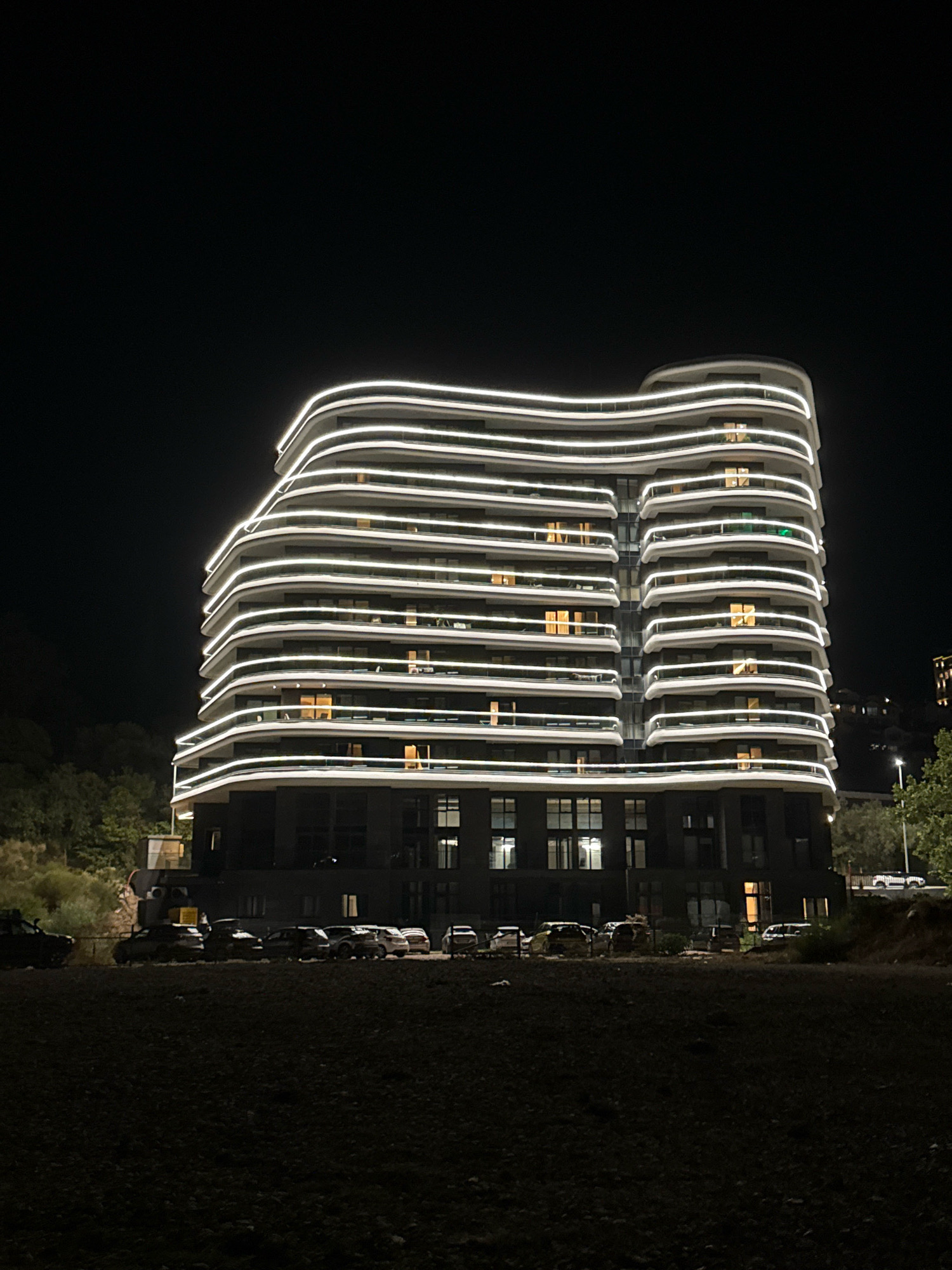 The image shows a modern, multi-story building with a distinctive curved facade illuminated by horizontal white LED strip lights at night. The building appears to be located in Boreti, Montenegro, as indicated by the context. There are several cars parked in a lot or street in front of the building. The environment is dark, with the exception of the artificial lighting from the building and some of the cars. Trees are visible in the background on the left side of the frame, and more of the surrounding landscape, possibly a hillside, is visible on the right. The overall scene suggests an urban or semi-urban setting at night.