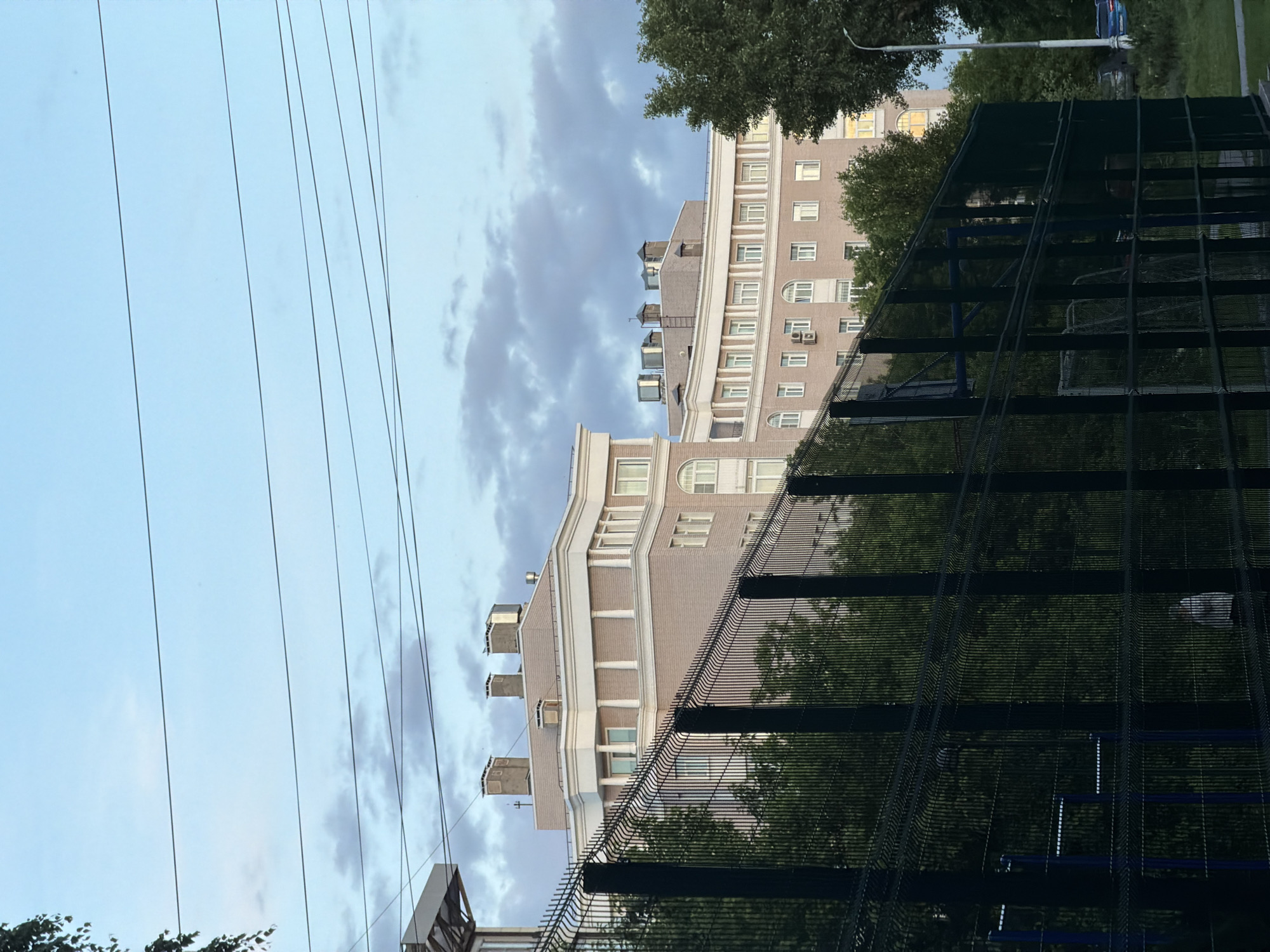 An outdoor urban scene in Moscow, Russia, captured from a low angle, looking upwards. The upper portion of the frame features a light blue sky with scattered greyish clouds. Several dark, thin utility wires stretch across the sky horizontally from left to right.
Below the sky, on the right side of the frame, stand multiple multi-story buildings. They are constructed with light brown brick or stone, featuring prominent white architectural details such as window frames, cornices, and decorative pillars on one structure. Numerous windows are visible, some with arched tops, and air conditioning units are mounted on the facades of one building. The roofs are dark, with several box-like structures, possibly vents or chimneys, protruding upwards. Green foliage from trees is visible in front of and behind the buildings.
In the foreground and mid-ground on the right, a tall, dark metal fence with a grid pattern encloses an area, likely a sports court. A blue structure, possibly part of a basketball hoop or goalpost, is discernible through the mesh of the fence. Further back, beyond the fence and trees, a dark blue car and a street light pole are partially visible. No human activity or visible text is present. The soft, diffuse lighting suggests an overcast day or late afternoon/early evening.