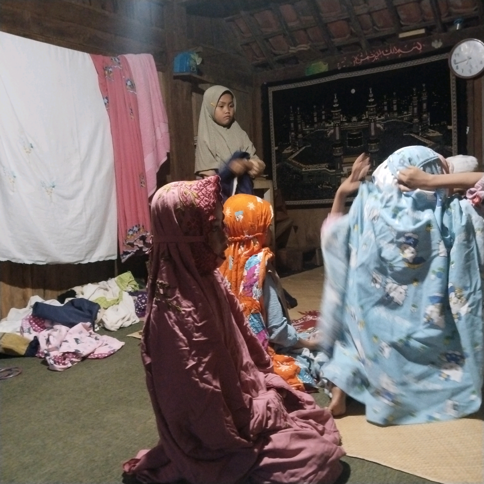 In a dimly lit room with a rustic wooden ceiling, four individuals, likely children, are engaged in activity on a green carpeted floor. Three of the individuals are wearing hijabs. The person on the far right is wearing a light blue hijab with a cartoon pattern, and their arms are raised, suggesting they are in the process of dressing or playing. To their left, another individual, facing away from the camera, wears a bright orange hijab with a patterned fabric. In the foreground, a third individual, also facing away, is draped in a deep pink hijab with intricate embroidered patterns. A fourth individual, partially visible in the background and to the left, is wearing a light brown hijab. The room is cluttered with various items, including folded fabrics of different colors and patterns, suggesting a domestic or communal space. A large framed tapestry depicting a cityscape, possibly Mecca, hangs on the wall behind the individuals, above which Arabic calligraphy is visible. A circular clock with Roman numerals is mounted on the wall to the right of the tapestry. The overall impression is a scene of everyday life within a modest dwelling in Banyubunih, Indonesia.