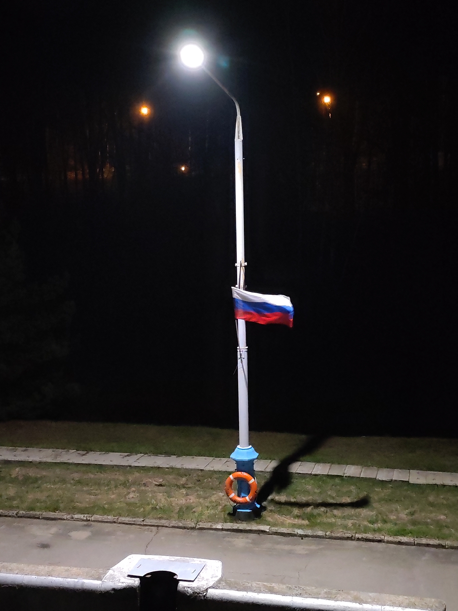 At night, a Russian flag flies from a white pole illuminated by a bright light at the top of the pole. An orange life preserver is attached to the base of the pole. The scene appears to be in a park-like setting, with a sidewalk and grass visible. Trees are present in the background.